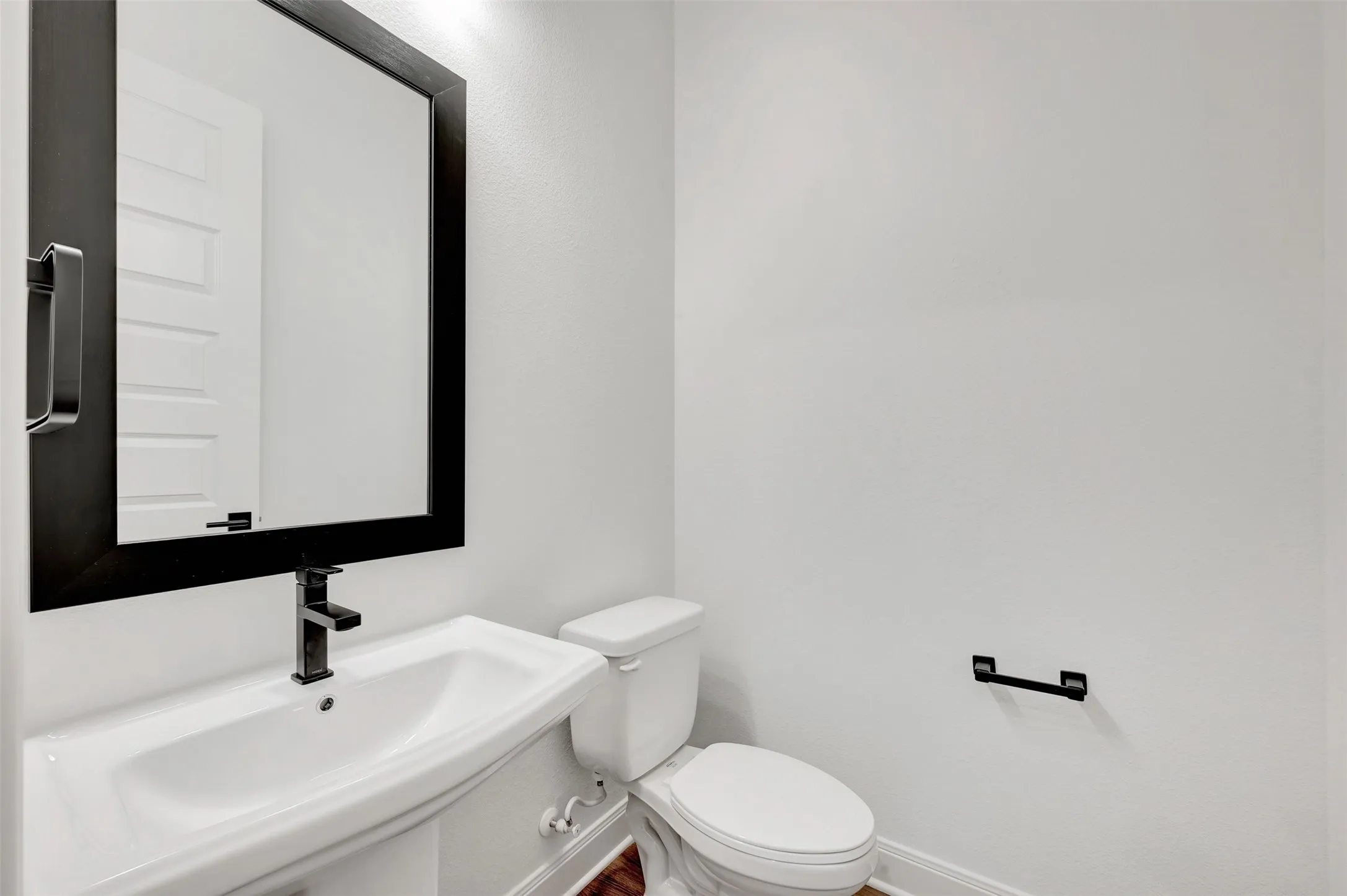 White pedestal sink with a matte black faucet, framed mirror, and wood-finish flooring