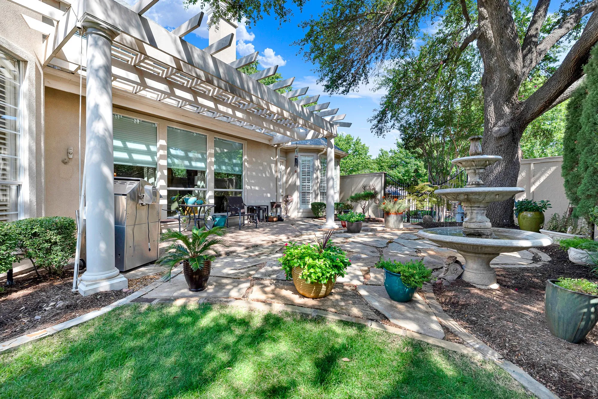 Classic white columns support an open-trellis pergola over a stone patio that transitions seamlessly to a grassy yard and fountain feature. It offers the ultimate setting for dining alfresco under the shade of magnificent mature trees.