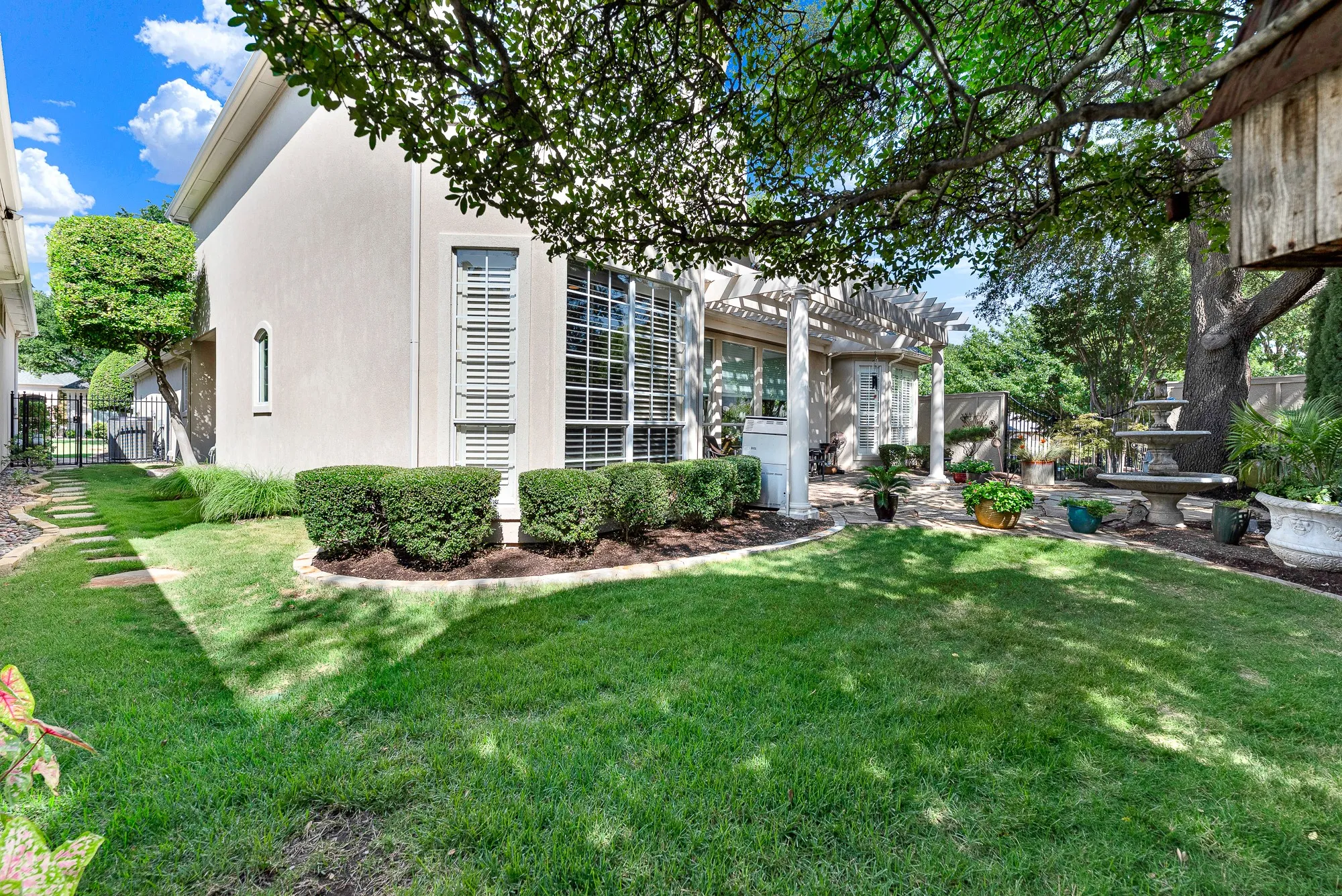A lush green lawn with stepping stones winds around manicured boxwood hedges toward the covered patio and stone fountain. Children and pets have plenty of soft grass to run and play safely in this fully fenced backyard space