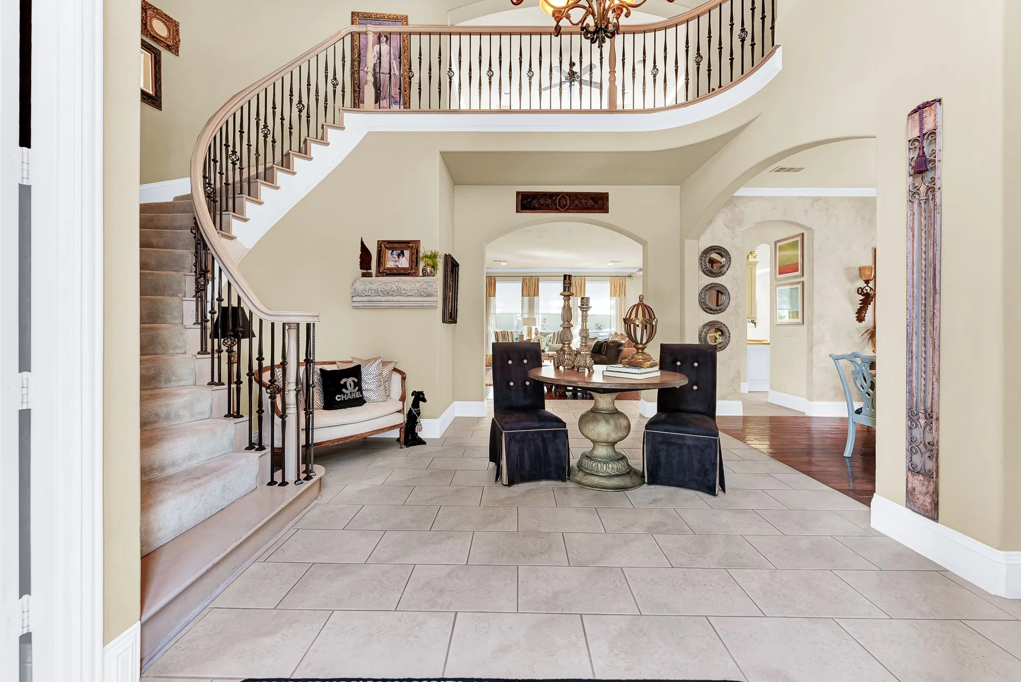 A dramatic curved staircase with ornate balusters leads to an open second-floor gallery overlooking neutral tile flooring and soft archways. It creates a stunning backdrop for hosting holiday gatherings and greeting guests in style.