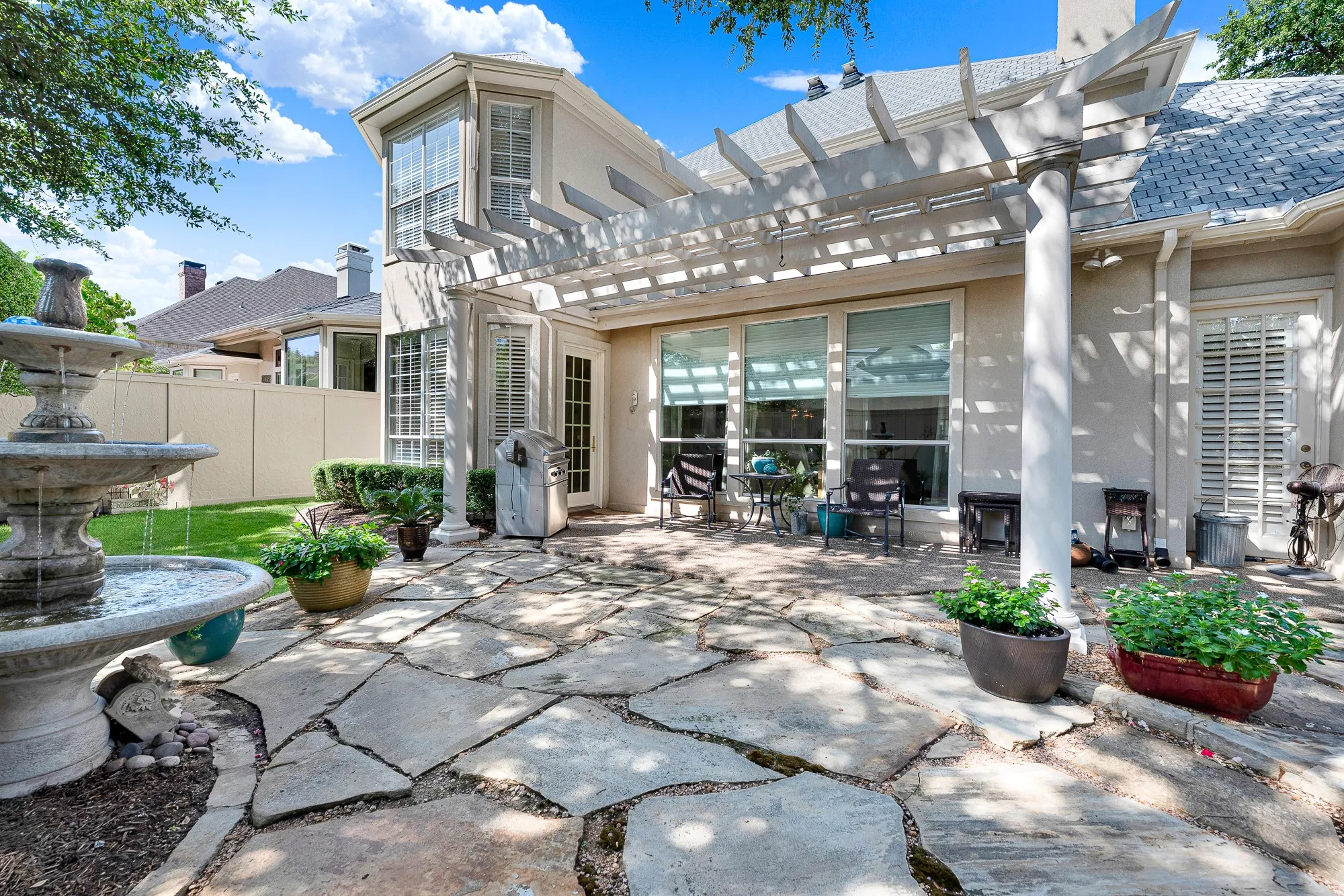 A multi-tiered stone fountain serves as the focal point alongside an expansive flagstone terrace and white columned pergola. Imagine unwinding outside with a cool drink on warm evenings in this serene outdoor sanctuary.