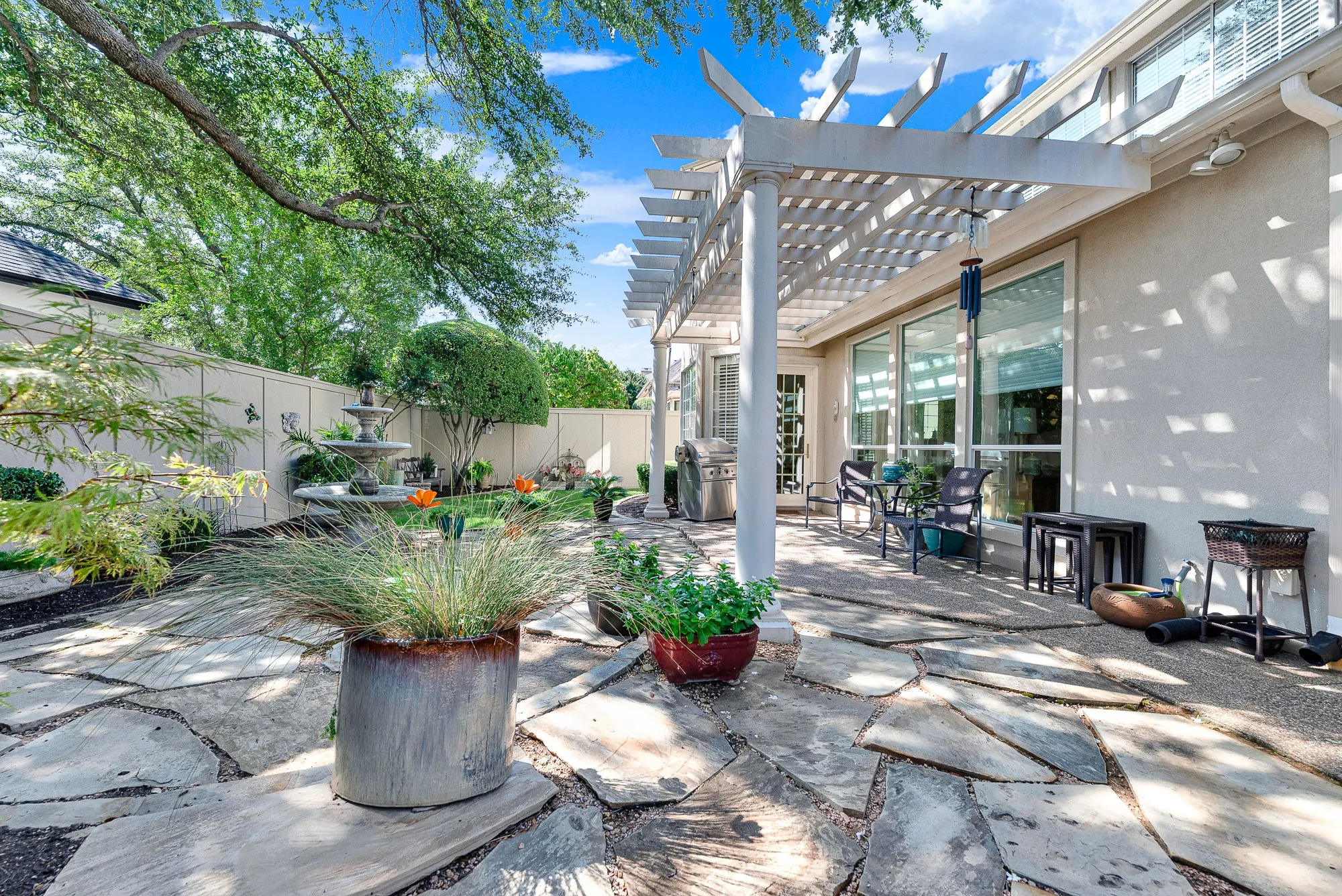 A spacious flagstone patio sits beneath a classic white pergola, framed by mature shade trees and a tiered stone fountain. It is a wonderful setting to host weekend barbecues or enjoy peaceful afternoons listening to gently cascading water.