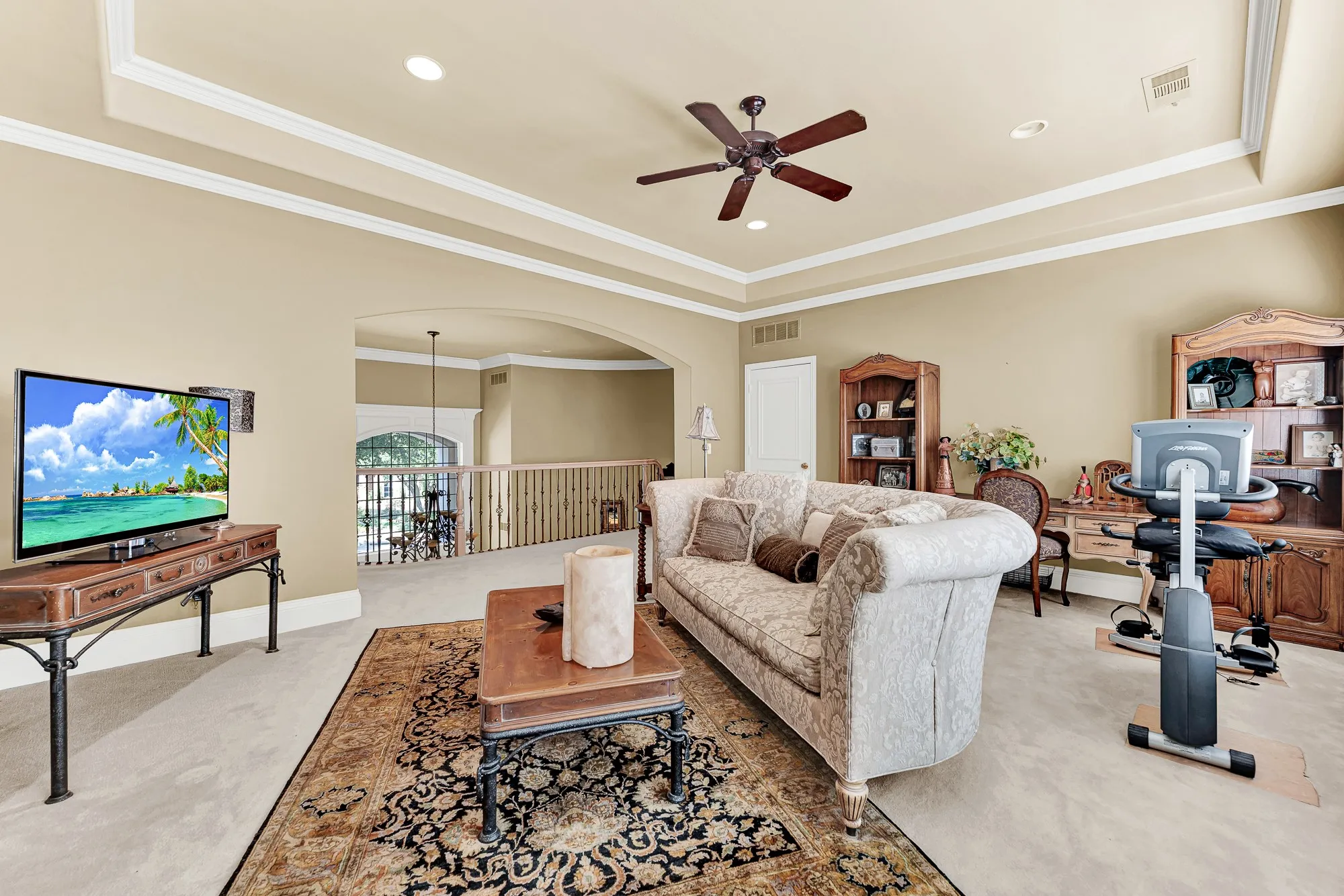 An arched opening with wrought-iron railing overlooks the lower level, highlighting tray ceilings, soft neutral walls, and plush flooring in this upstairs flex room. Hosting sleepovers or casual hangouts is effortless with this seamless connection to the rest of the home.