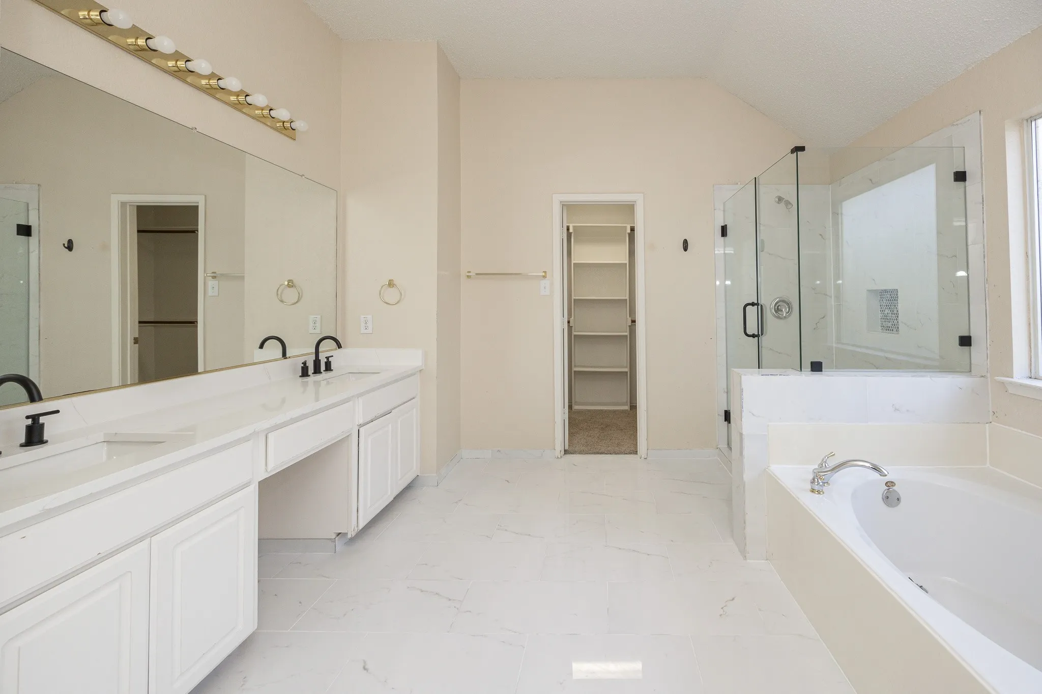 Dual vanity with white cabinetry and black fixtures, expansive mirror, separate glass-enclosed shower, built-in soaking tub, and vaulted ceiling