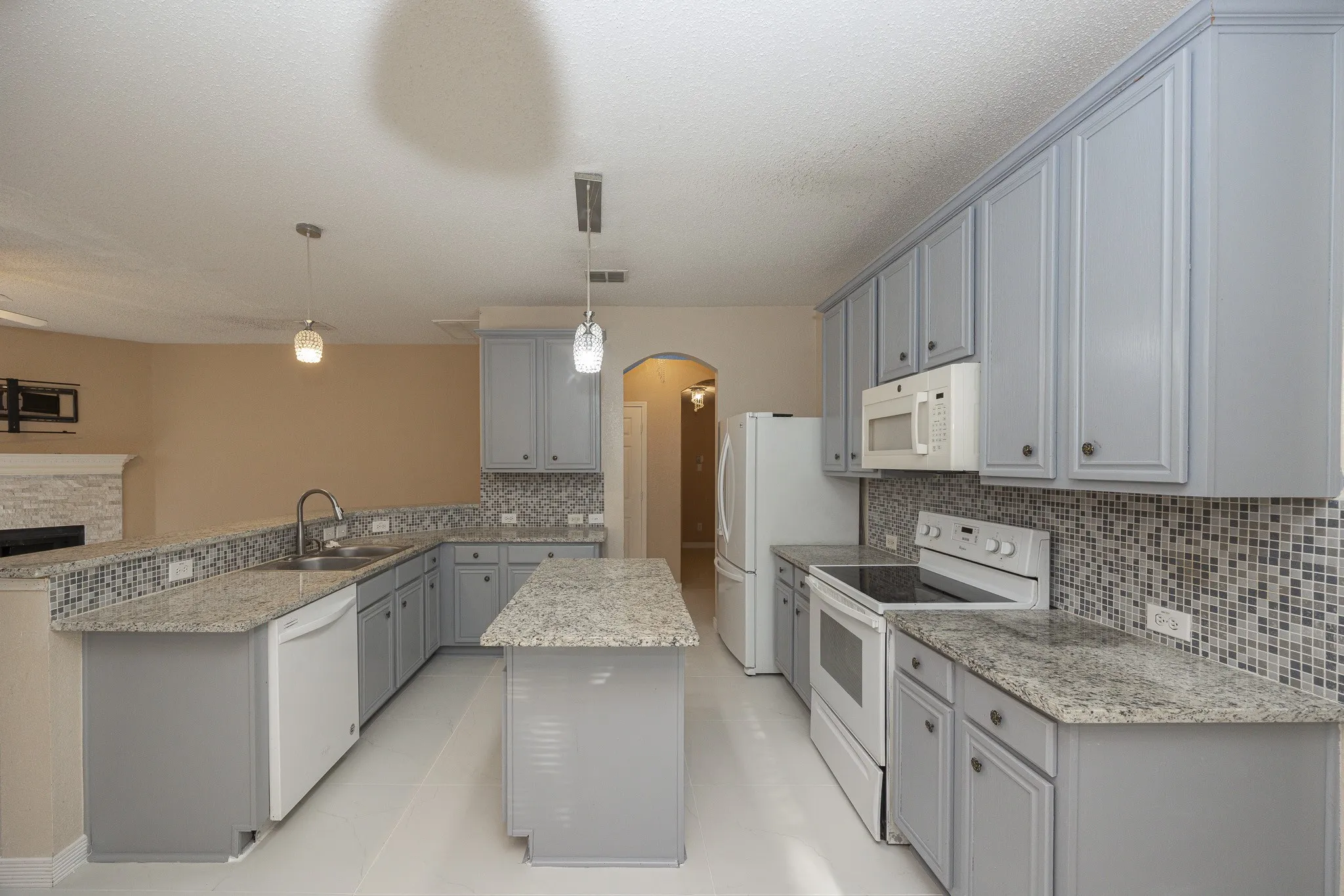 Kitchen featuring light gray cabinetry, granite countertops, mosaic tile backsplash, white appliances, and a central island