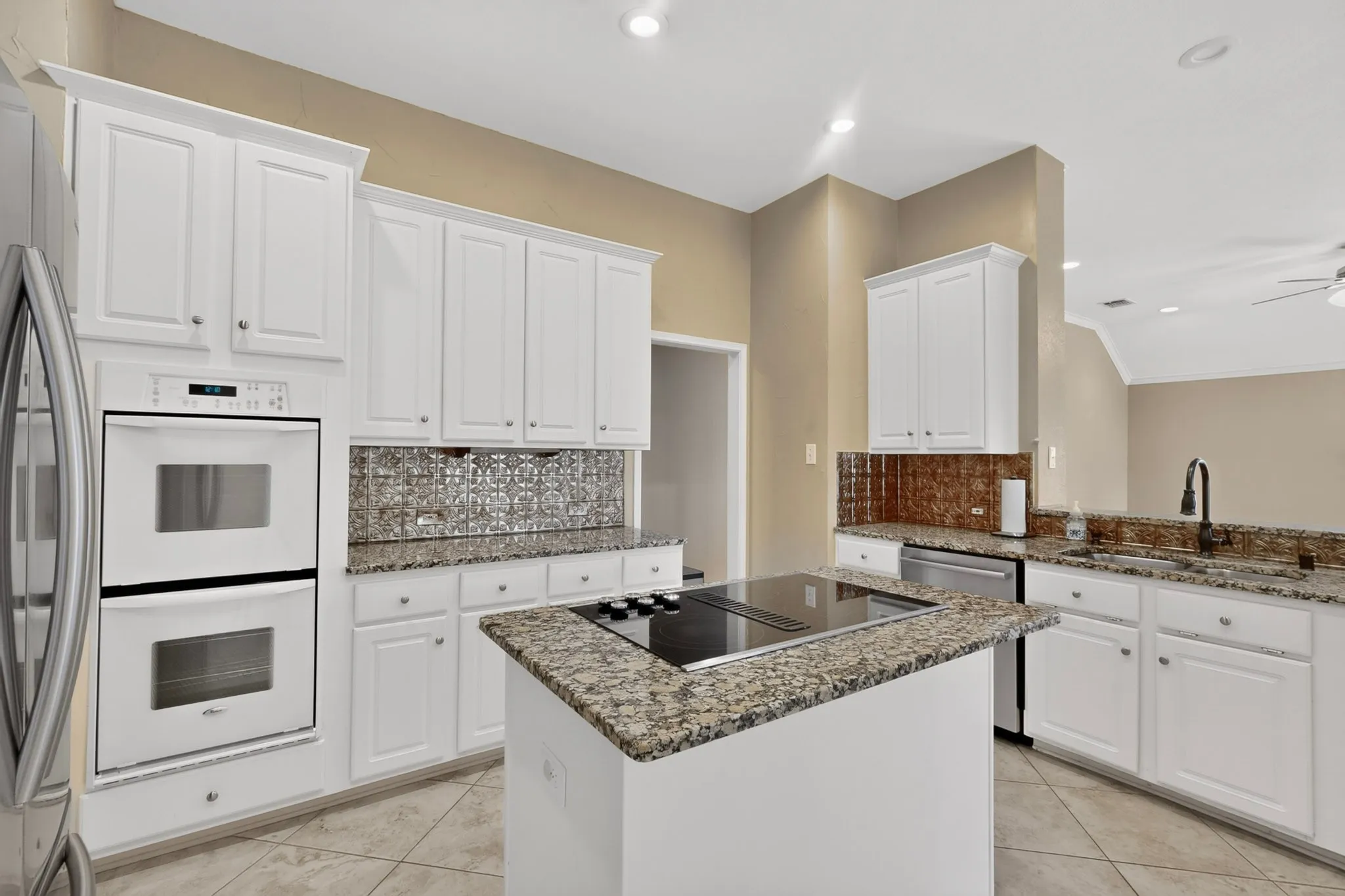 White cabinetry, granite countertops, tile flooring, built-in double ovens, and a kitchen island with an integrated cooktop