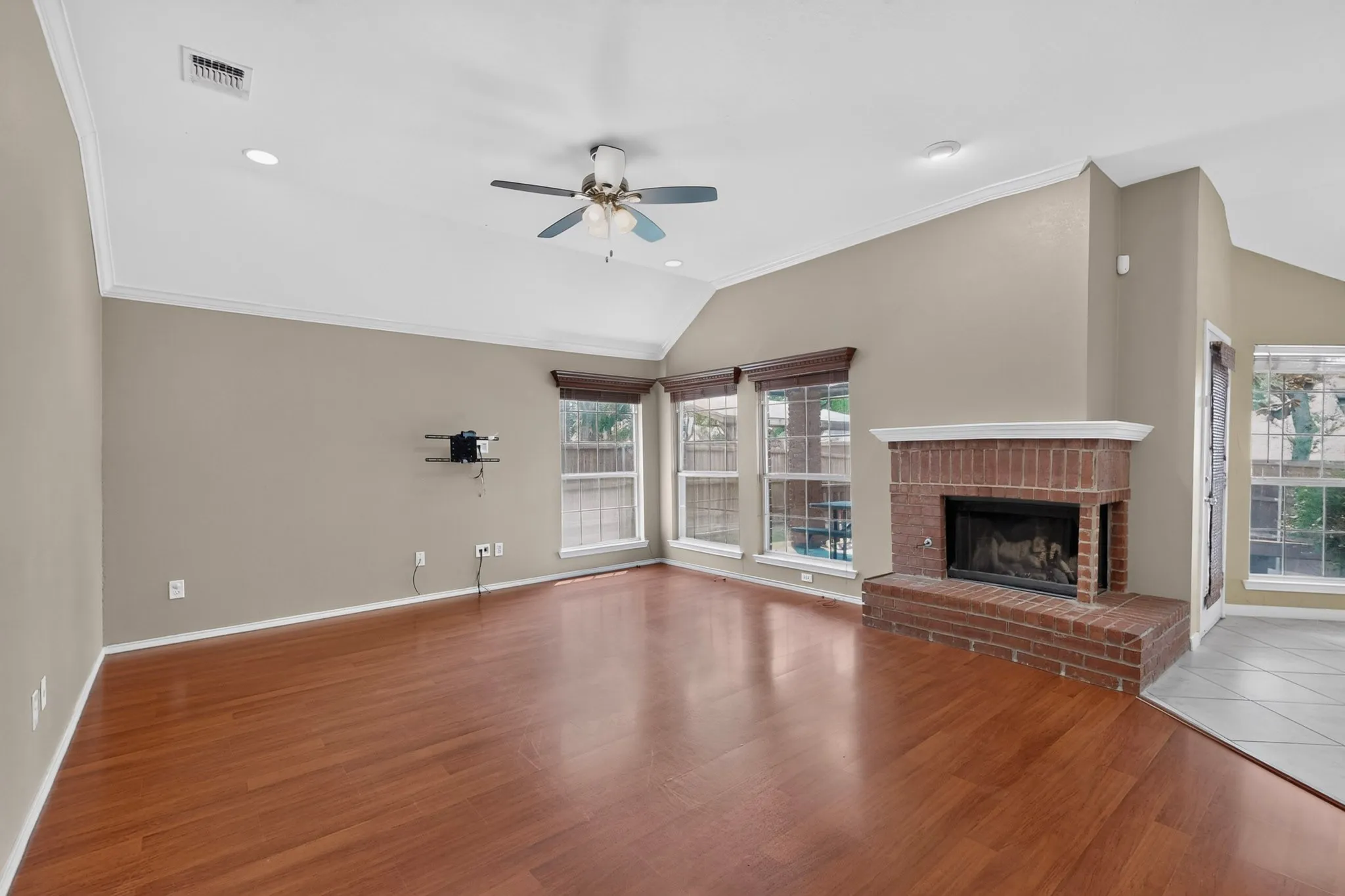 Spacious living area featuring wood-finish flooring, a brick fireplace with a white mantel, multiple windows, and a ceiling fan