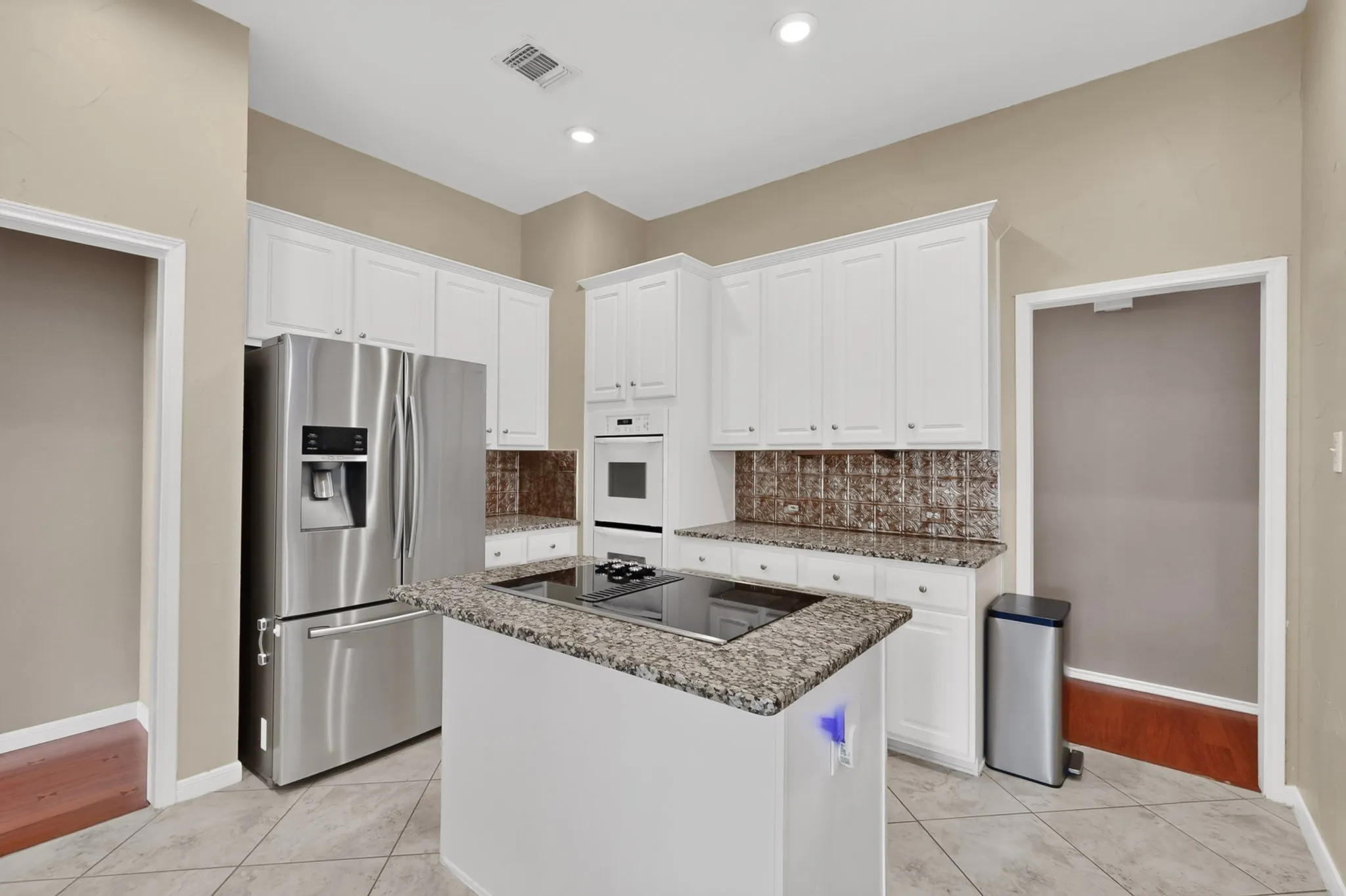 Kitchen featuring an island with a built-in cooktop, white upper and lower cabinetry, stainless steel refrigerator, built-in oven, and recessed lighting