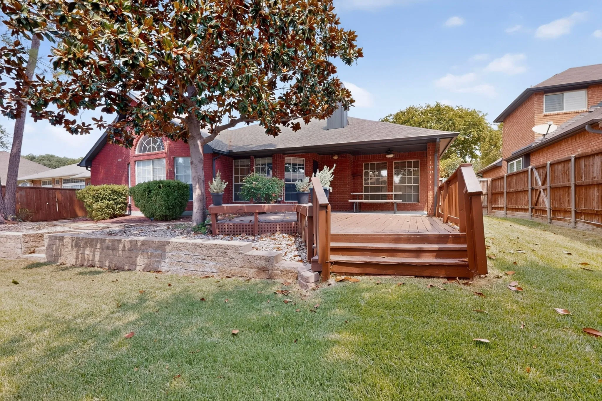 Rear exterior featuring a brick facade, covered patio, wooden deck with stairs, established tree, and a stone retaining wall