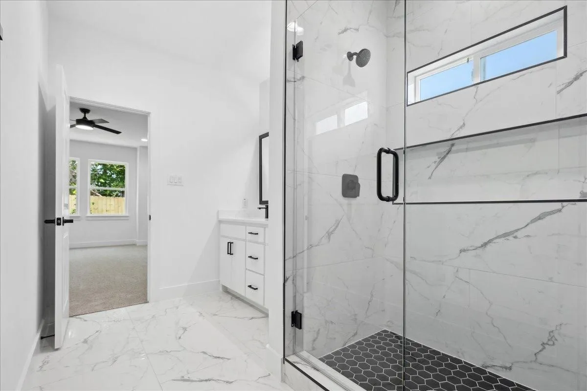Spa-inspired primary bathroom showcasing a frameless glass-enclosed shower with a linear rainfall showerhead, marble-look tile walls and flooring, a floating vanity with integrated storage, and a high transom window providing natural light and ventilation