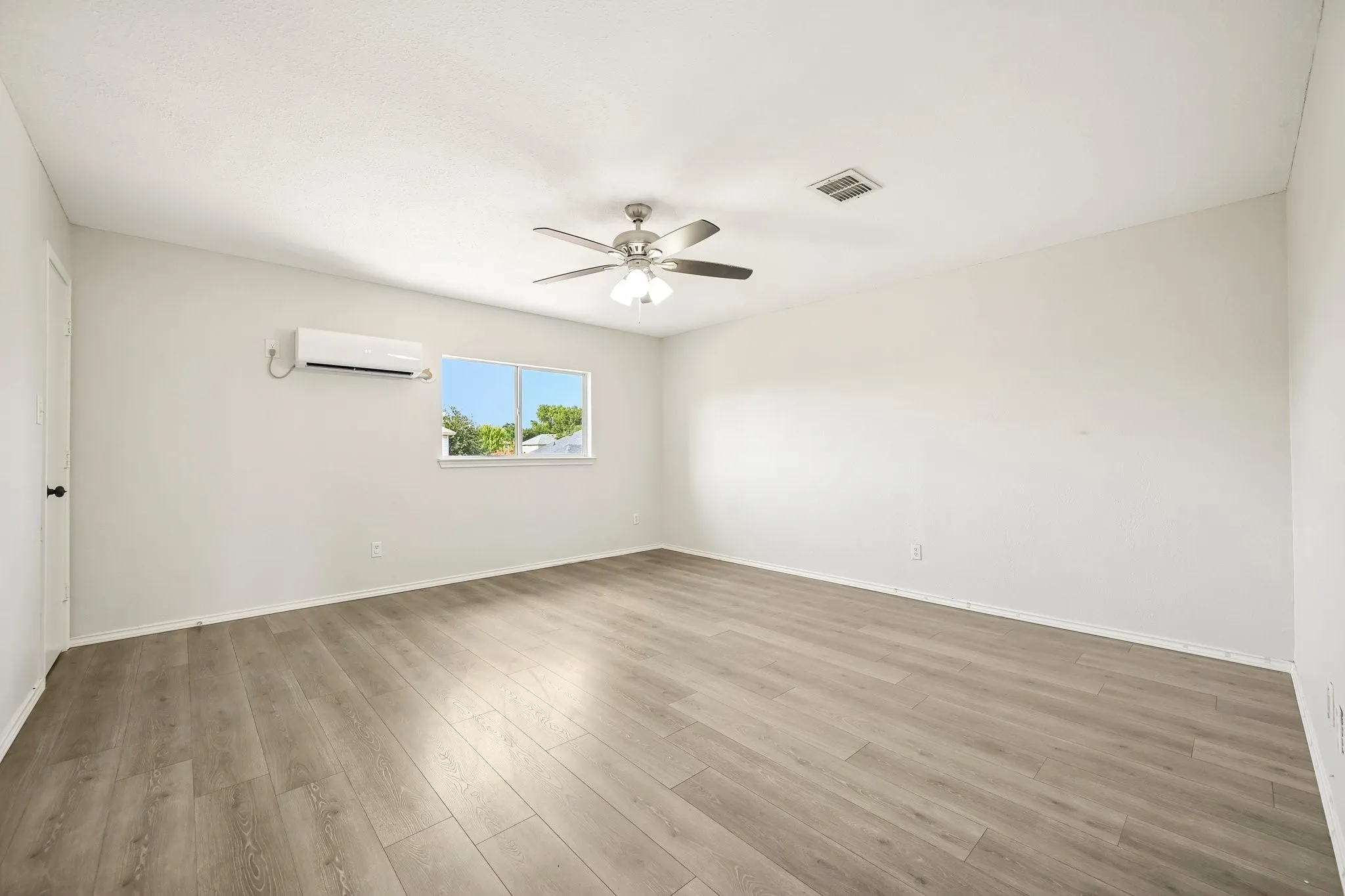 Wood-finish flooring, white walls, ceiling-mounted fan with integrated lighting, window with white trim, and a wall-mounted mini-split AC unit