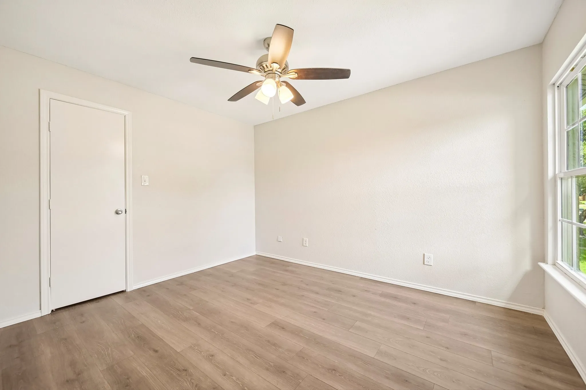 Wood-finish flooring, ceiling fan with integrated lighting, white interior door, and a multi-pane window