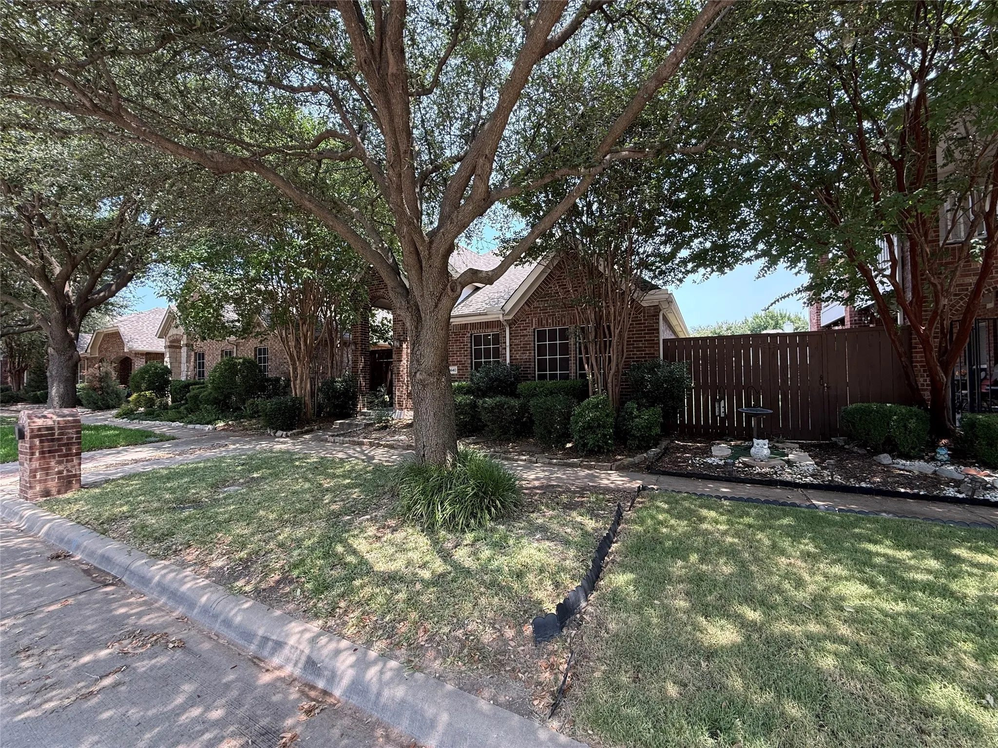 Mature trees providing ample shade, brick exterior with gabled roofline, front yard with established landscaping, and a privacy fence