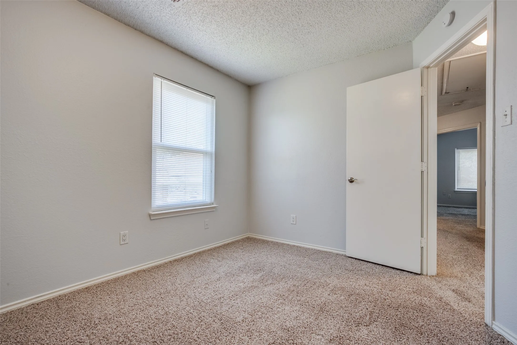 Neutral wall color with textured ceiling, bright window with blinds, and light-colored carpeting