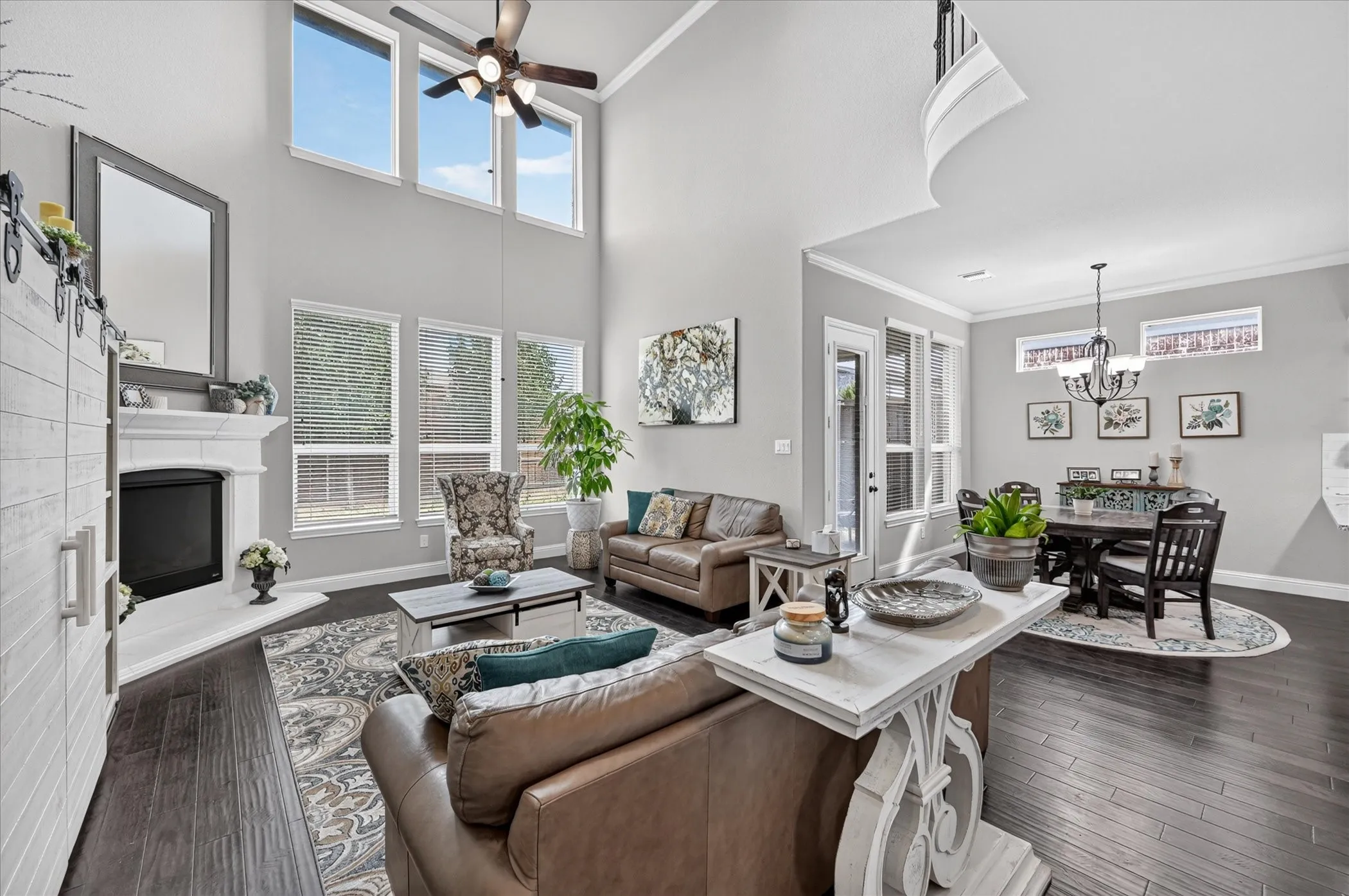 Spacious open-concept living area featuring soaring ceilings with ceiling fan, abundant natural light from multiple windows, dark wood-finish flooring, built-in fireplace with white surround, and coordinating dining space with pendant lighting and black wood chairs