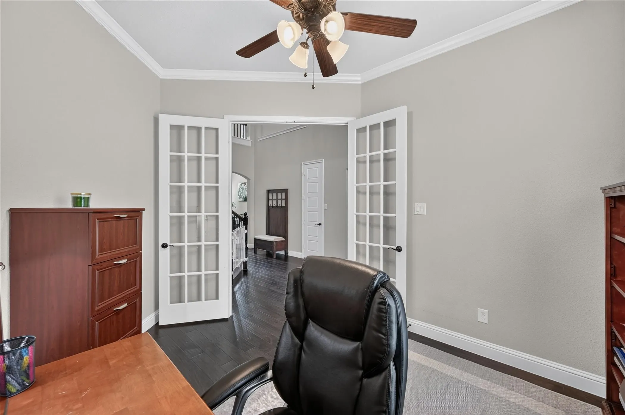 Professional home office featuring a spacious layout with dual French doors, wood-finish desk, ceiling fan with integrated lighting, and neutral wall finishes complemented by rich wood-tone cabinetry