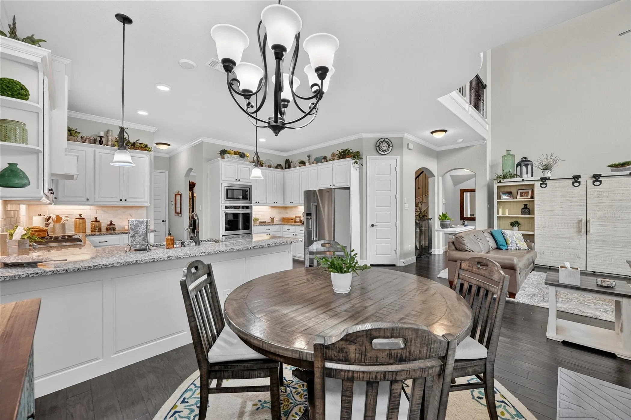 Open-concept kitchen and dining area featuring white cabinetry with glass-front upper cabinets, stainless steel appliances, granite countertops, wood-finish dining table, and a black chandelier with frosted glass shades suspended from a coffered ceiling