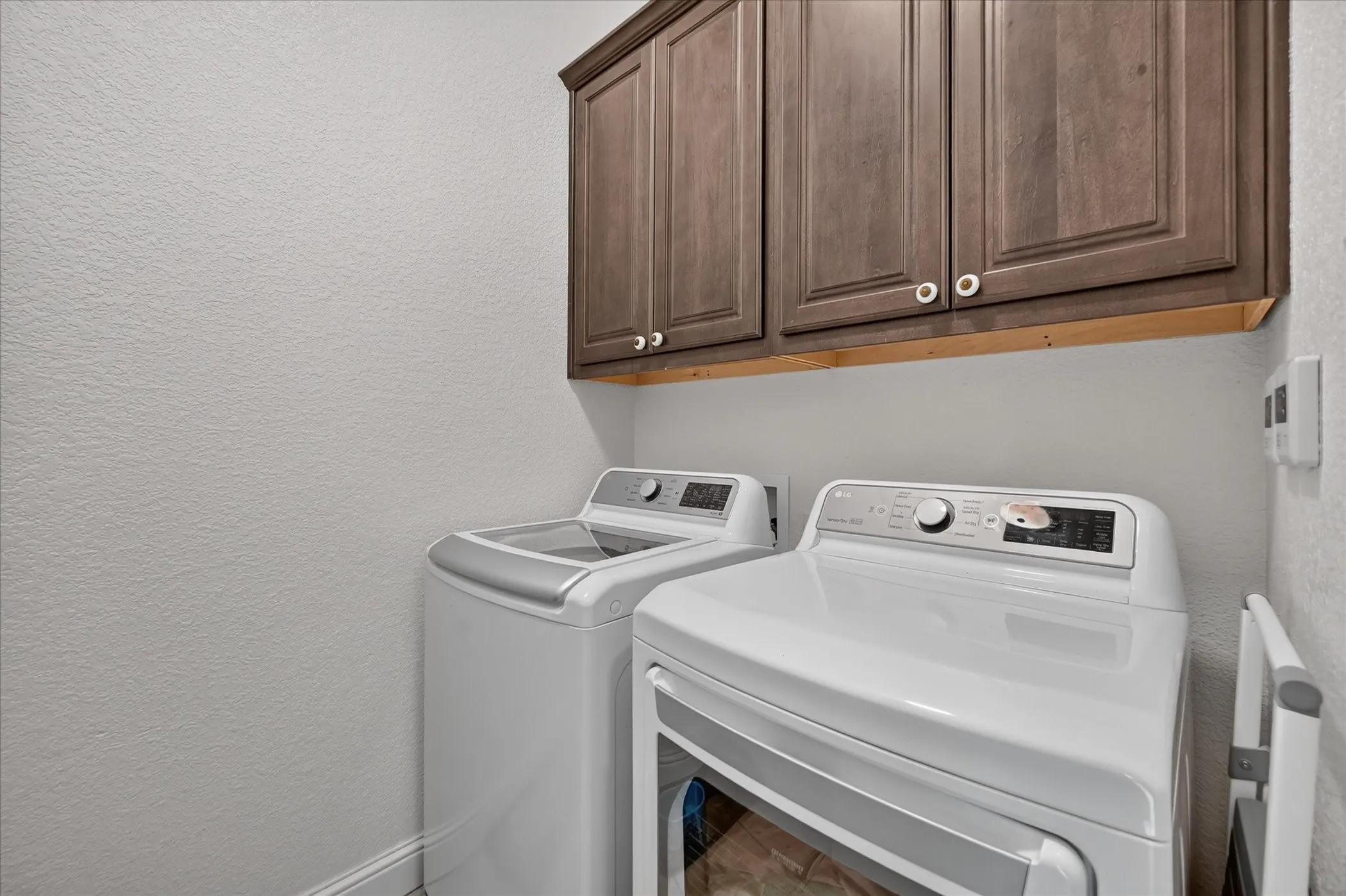 Dedicated laundry room featuring side-by-side white appliances with top-load and front-load configurations, wood-finish cabinetry with ample storage above, and neutral wall finishes for a clean, functional space