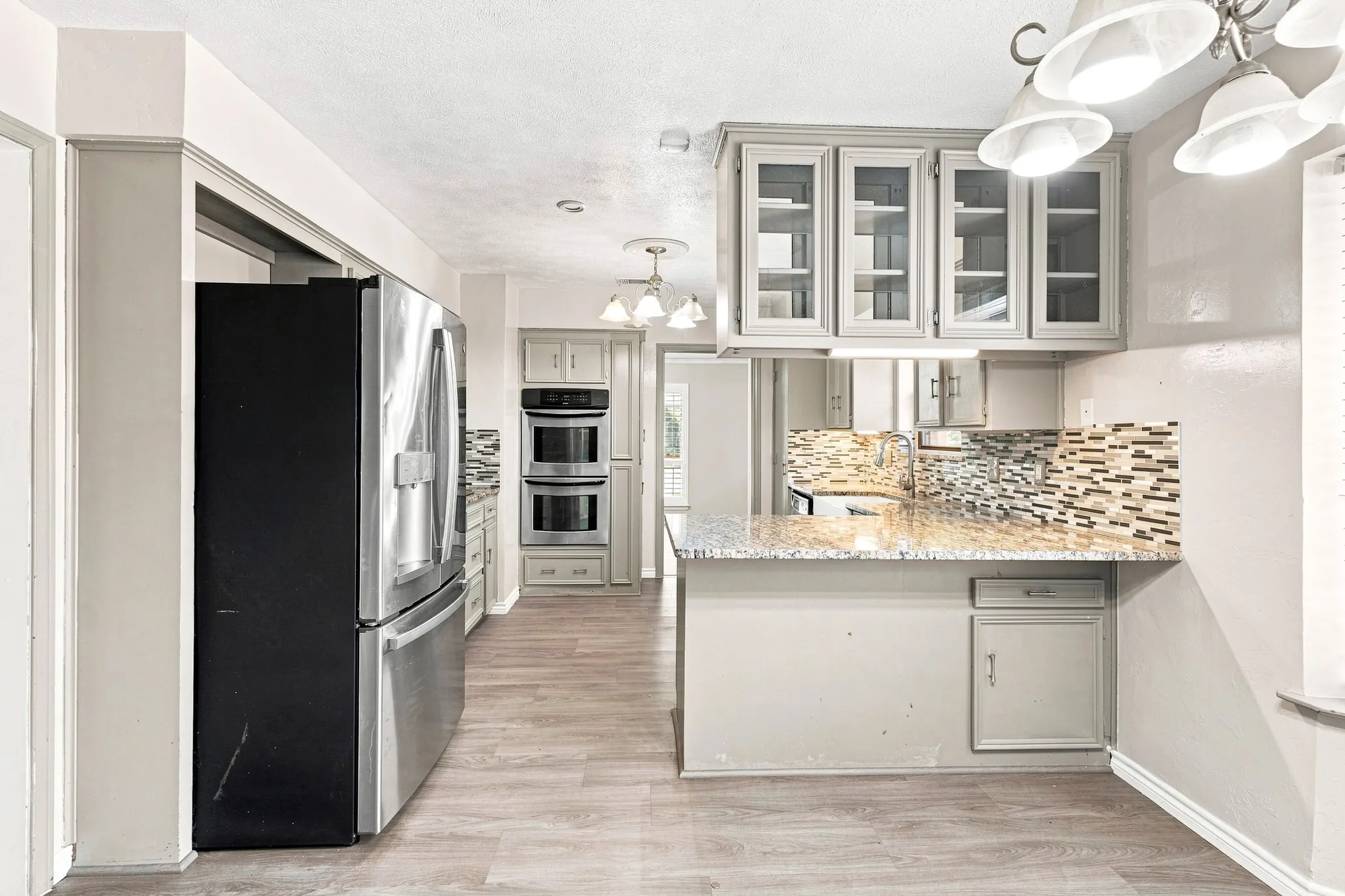 Stainless steel refrigerator, double wall ovens, wood-finish flooring, light grey cabinetry with glass inserts, and a patterned tile backsplash