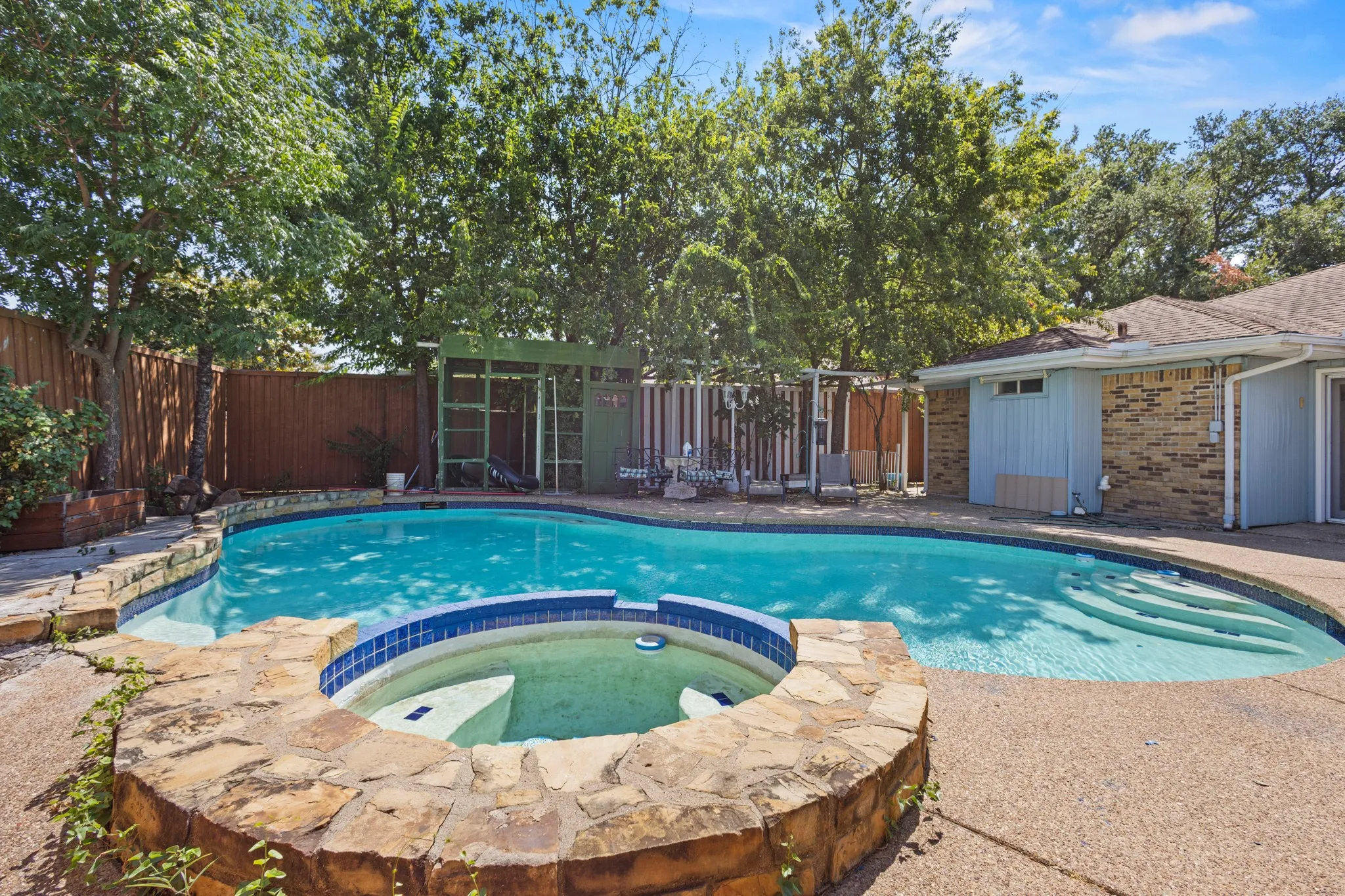 Kidney-shaped swimming pool with integrated spa featuring stone surround, concrete decking, and mature perimeter trees