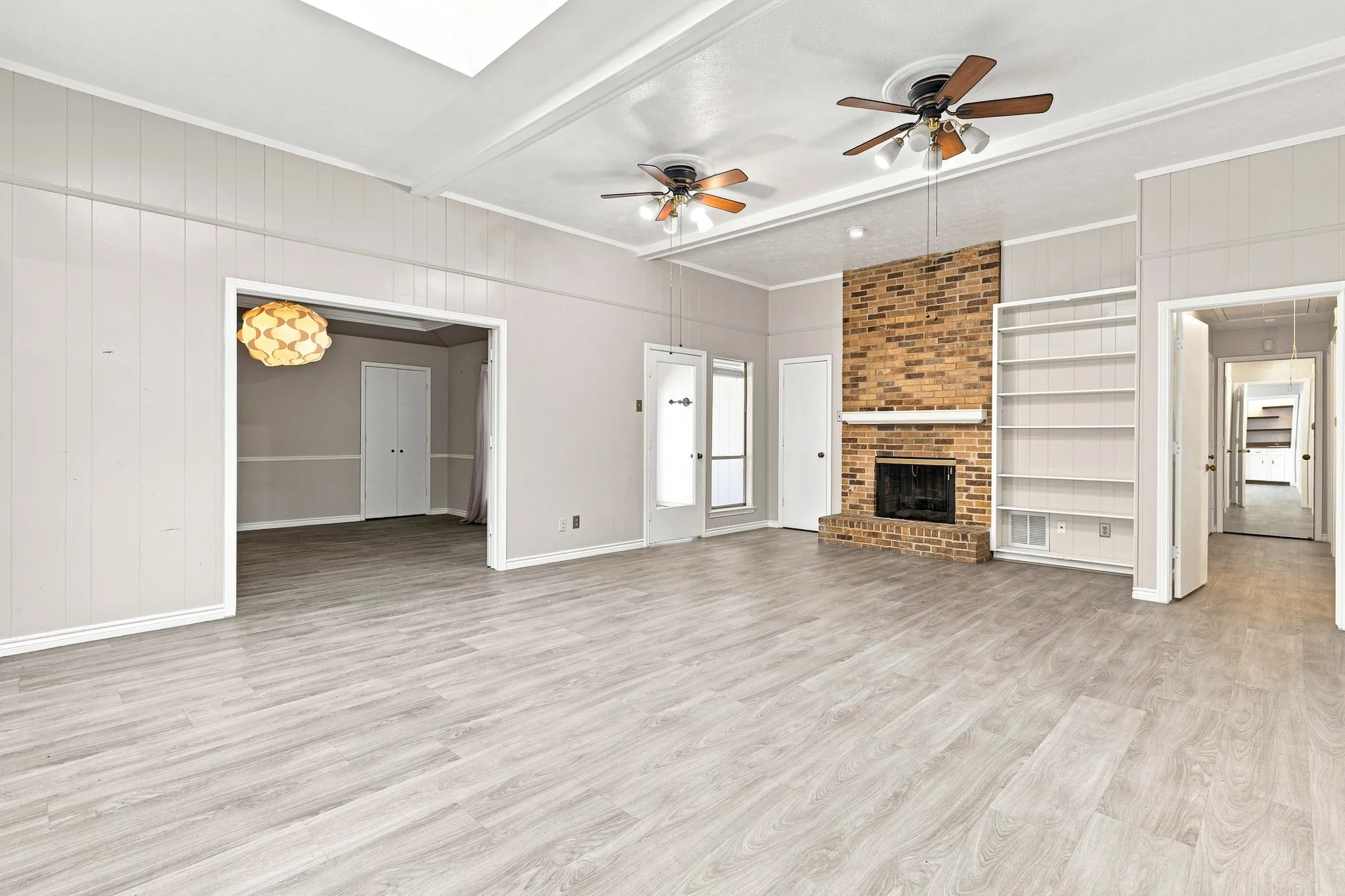 Expansive living area featuring light wood-finish flooring, a brick fireplace, built-in shelving, and two ceiling fans