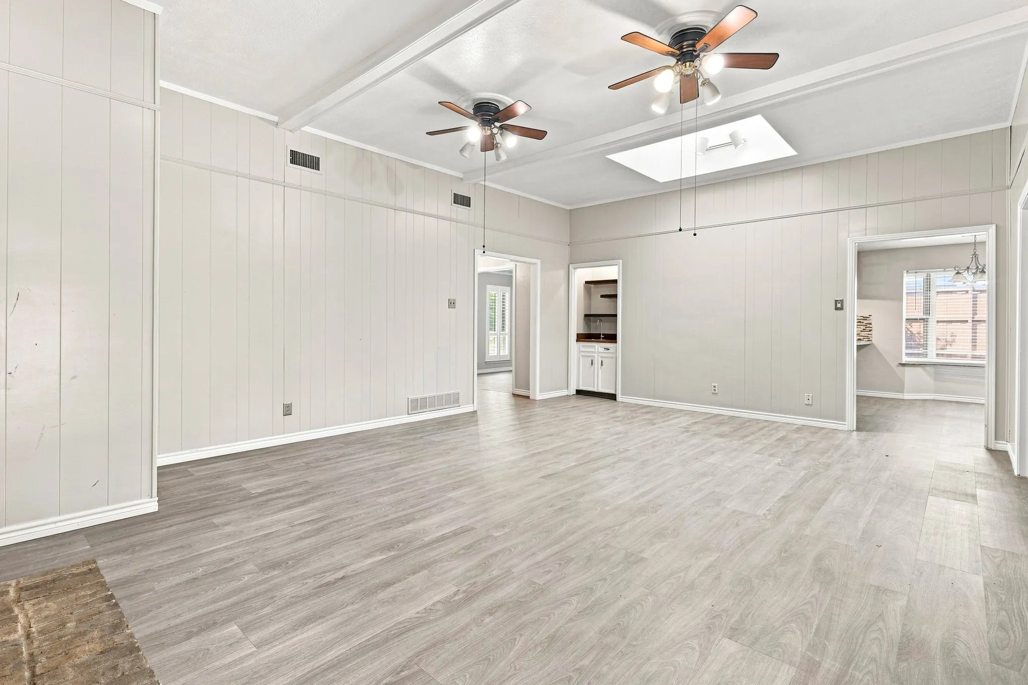 Spacious room featuring wood-finish flooring, vertical paneling, exposed ceiling beams, two ceiling fans, and a skylight
