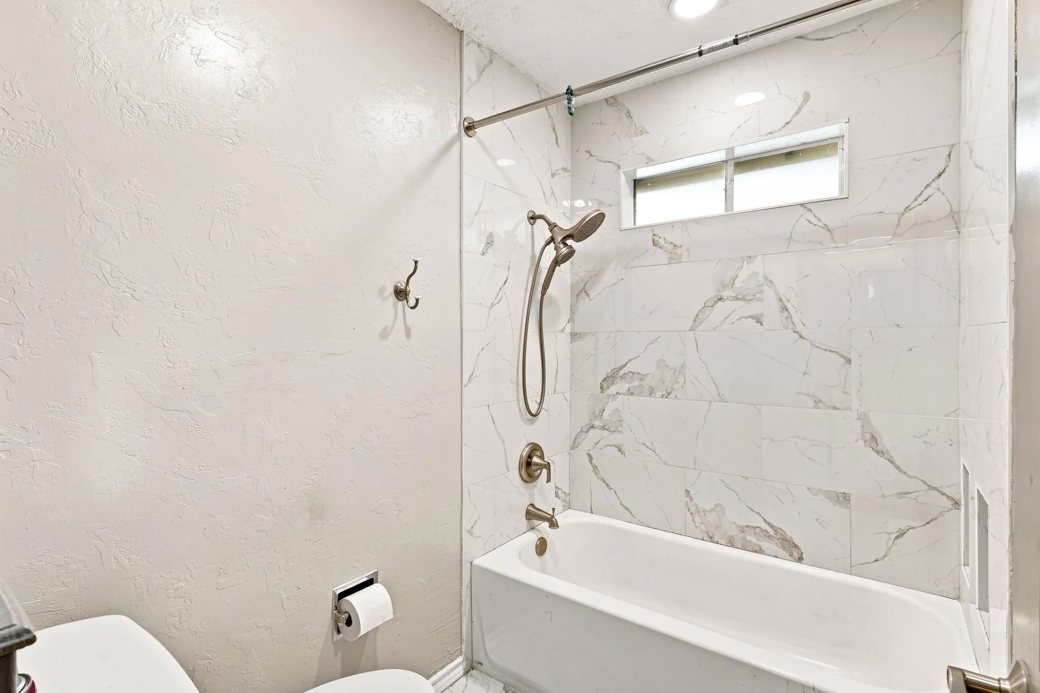 White textured wall finish, white tiled shower surround, recessed shower niche, brushed nickel shower fixtures, and a horizontal transom window