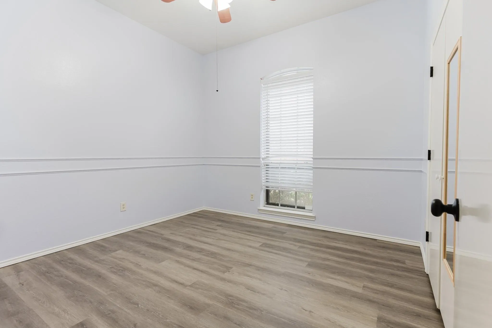 Wood-finish flooring, accent wall molding, arched window with blinds, ceiling fan, and paneled interior door with black hardware