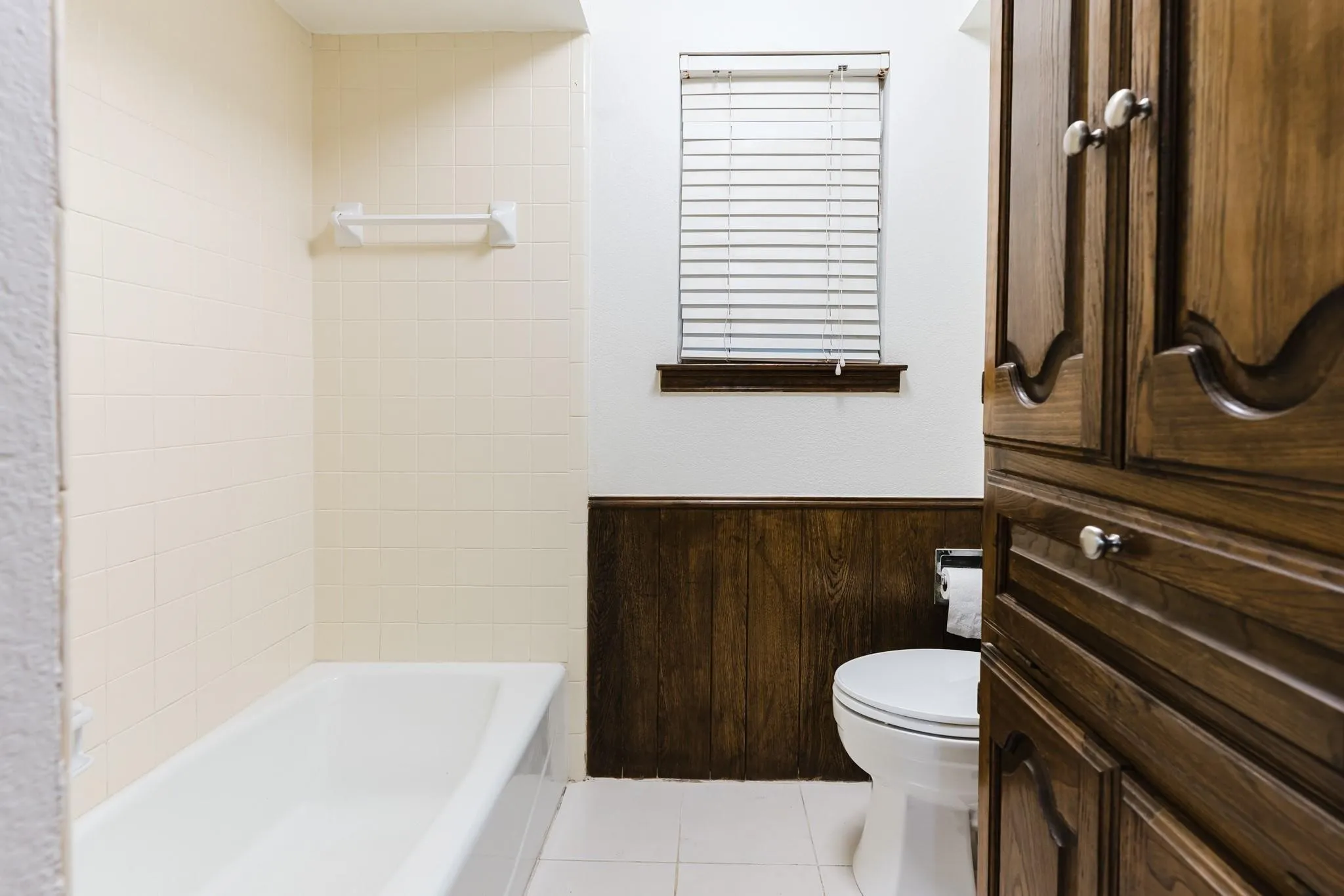 White ceramic tile flooring, wood paneling, built-in wooden cabinetry, white bathtub, and a window with blinds