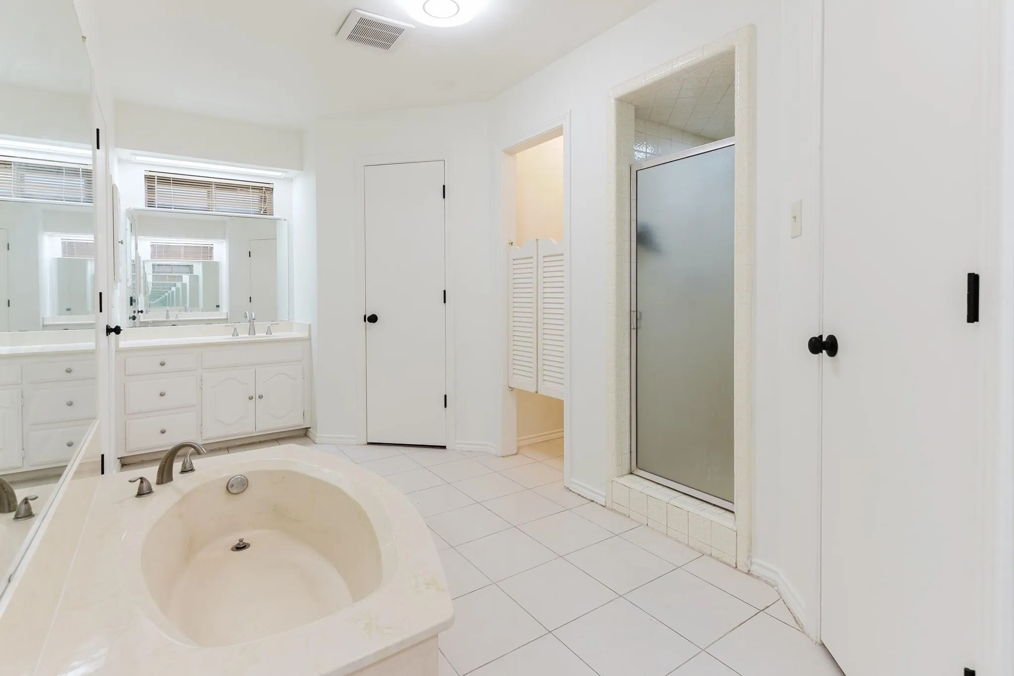 Built-in tub, dual vanity with undermount sinks, glass-enclosed shower, white tile flooring, and louvered bifold doors