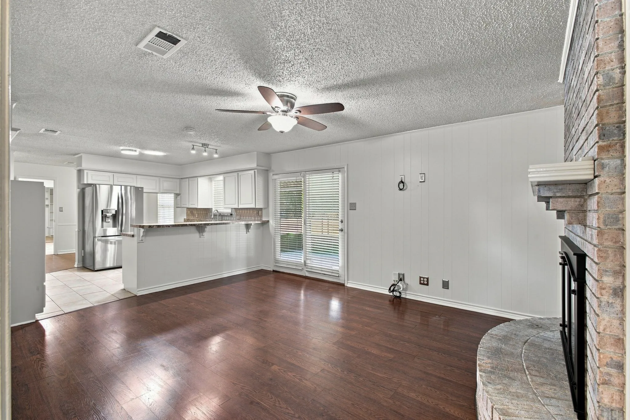 Wood-finish flooring, brick fireplace, white paneled wall, ceiling fan, and glass door to the backyard