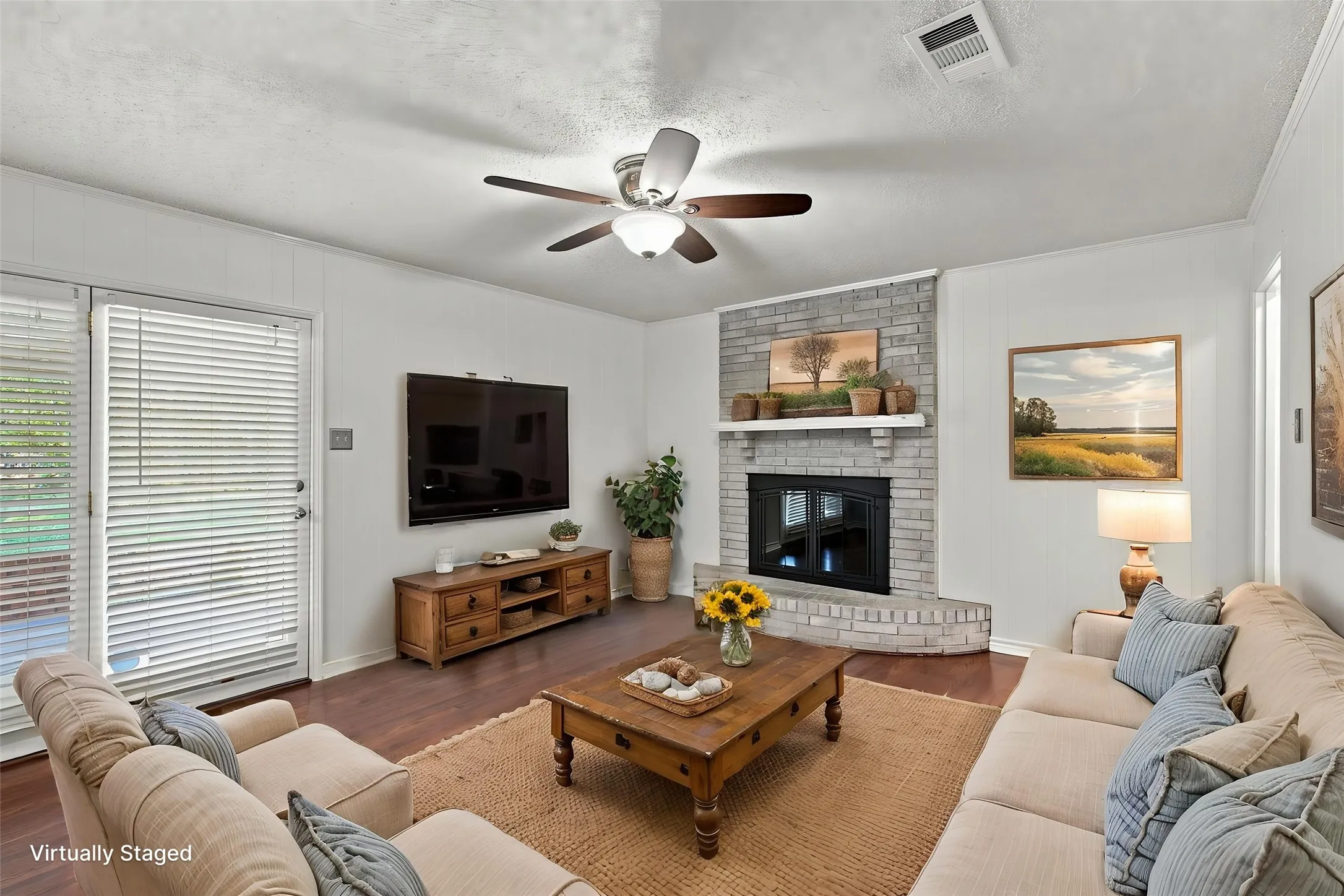 Wood-finish flooring, brick fireplace, white paneled wall, ceiling fan, and glass door to the backyard