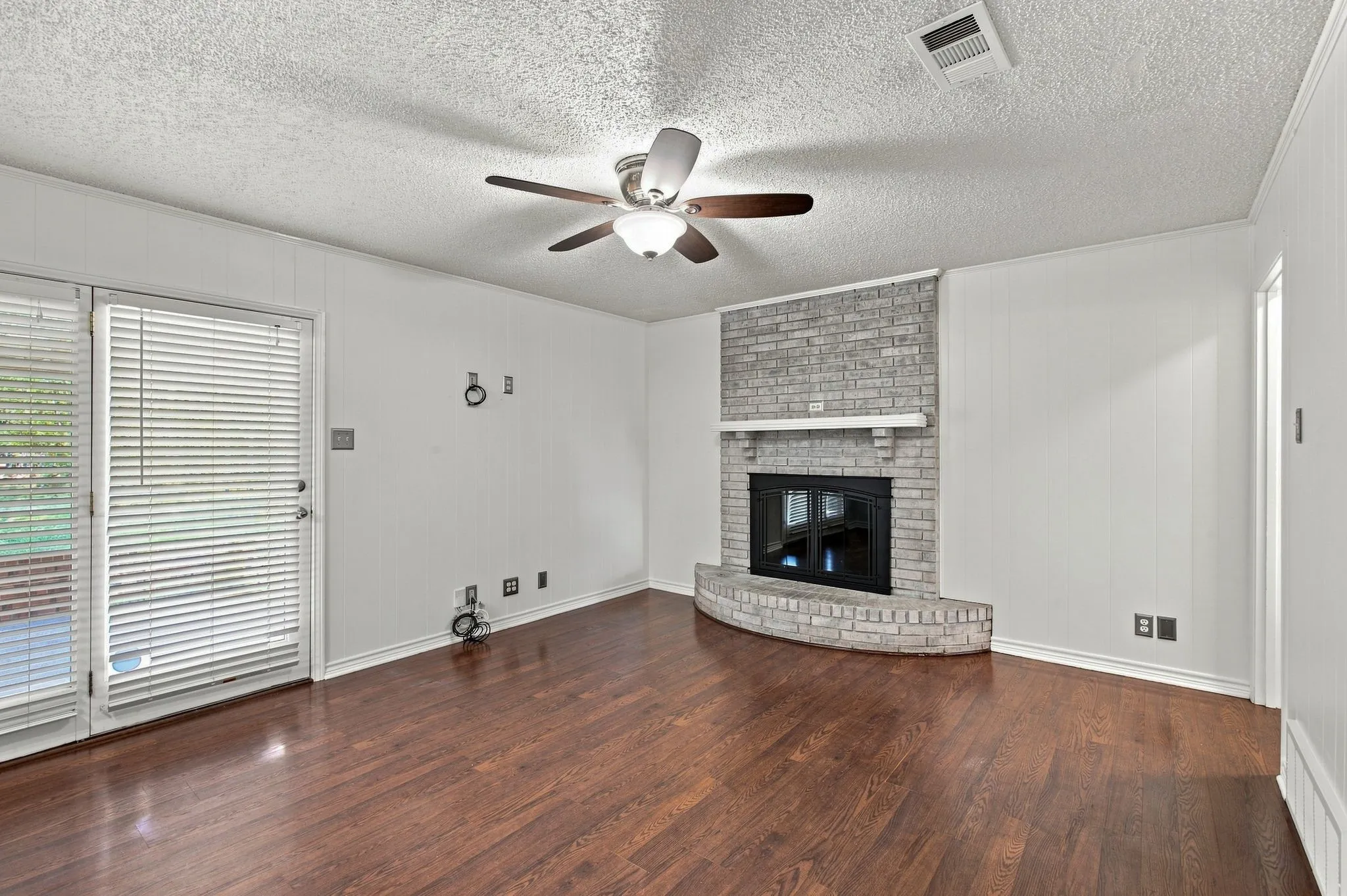 Wood-finish flooring, brick fireplace, white paneled wall, ceiling fan, and glass door to the backyard