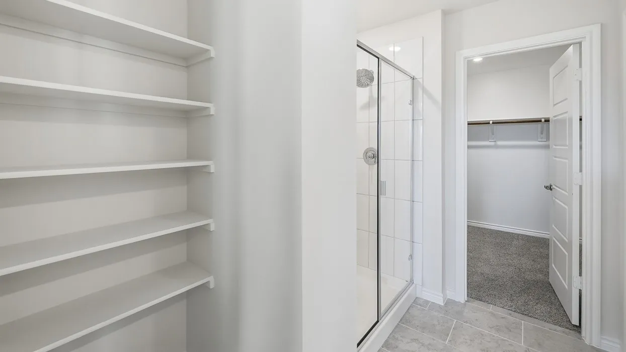 Built-in shelving, glass-enclosed shower with tile surround, and an open doorway leading to a carpeted space with closet rod and shelf