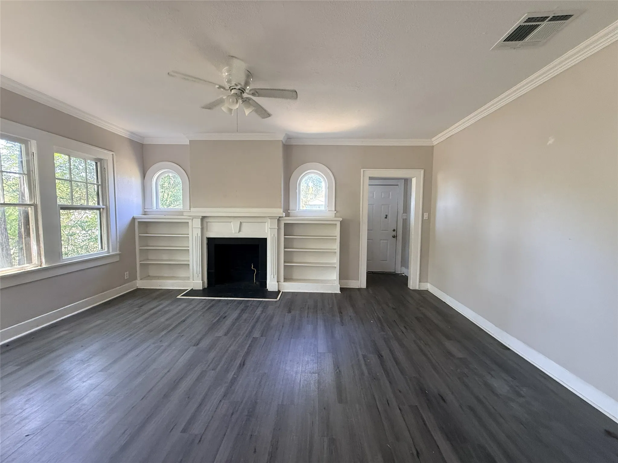 Spacious room featuring wood-finish flooring, a decorative fireplace with mantel, built-in shelving, two windows, and crown molding