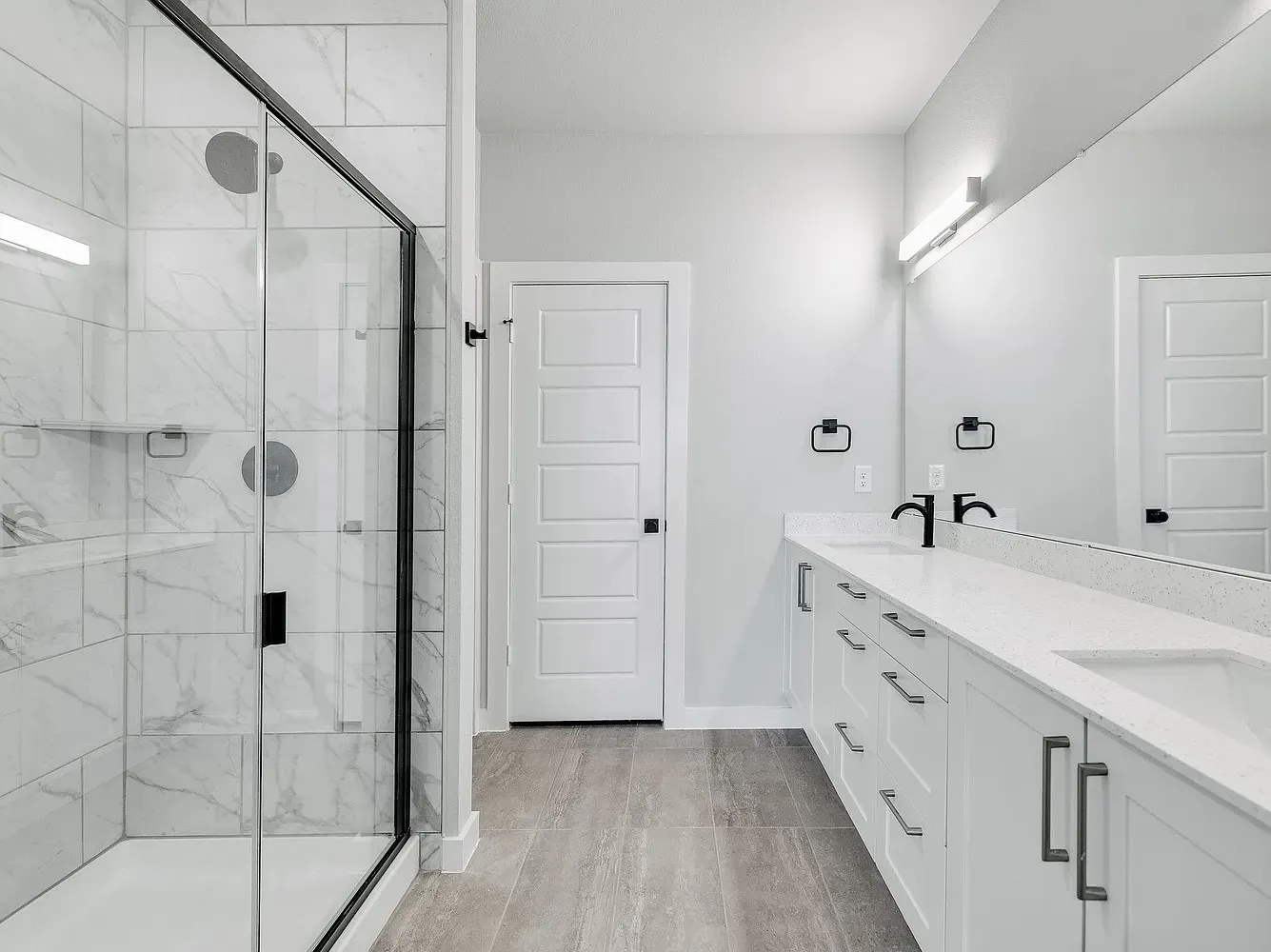 Spacious bathroom featuring a glass-enclosed shower with white tiled walls, a dual vanity with white cabinetry, integrated sinks, and matte black fixtures
