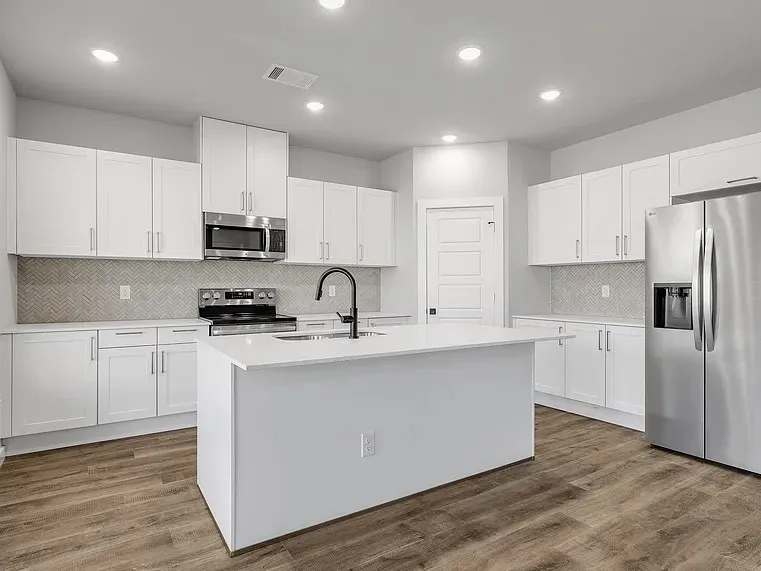Spacious kitchen featuring white cabinetry, a center island with sink, stainless steel appliances, herringbone tile backsplash, and wood-finish flooring