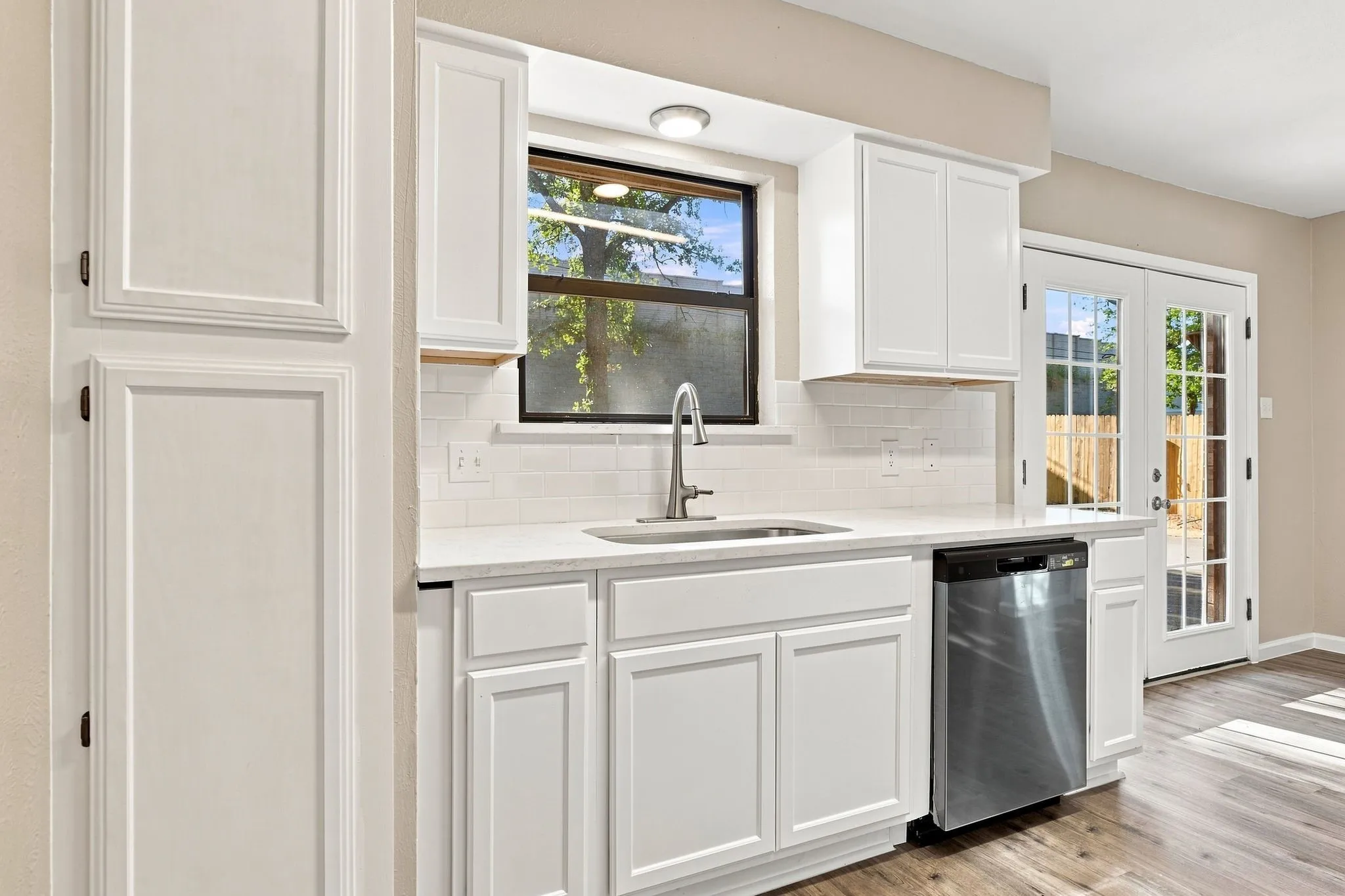 Wood-finish flooring, white cabinetry, integrated dishwasher, white subway tile backsplash, and double glass-paneled doors