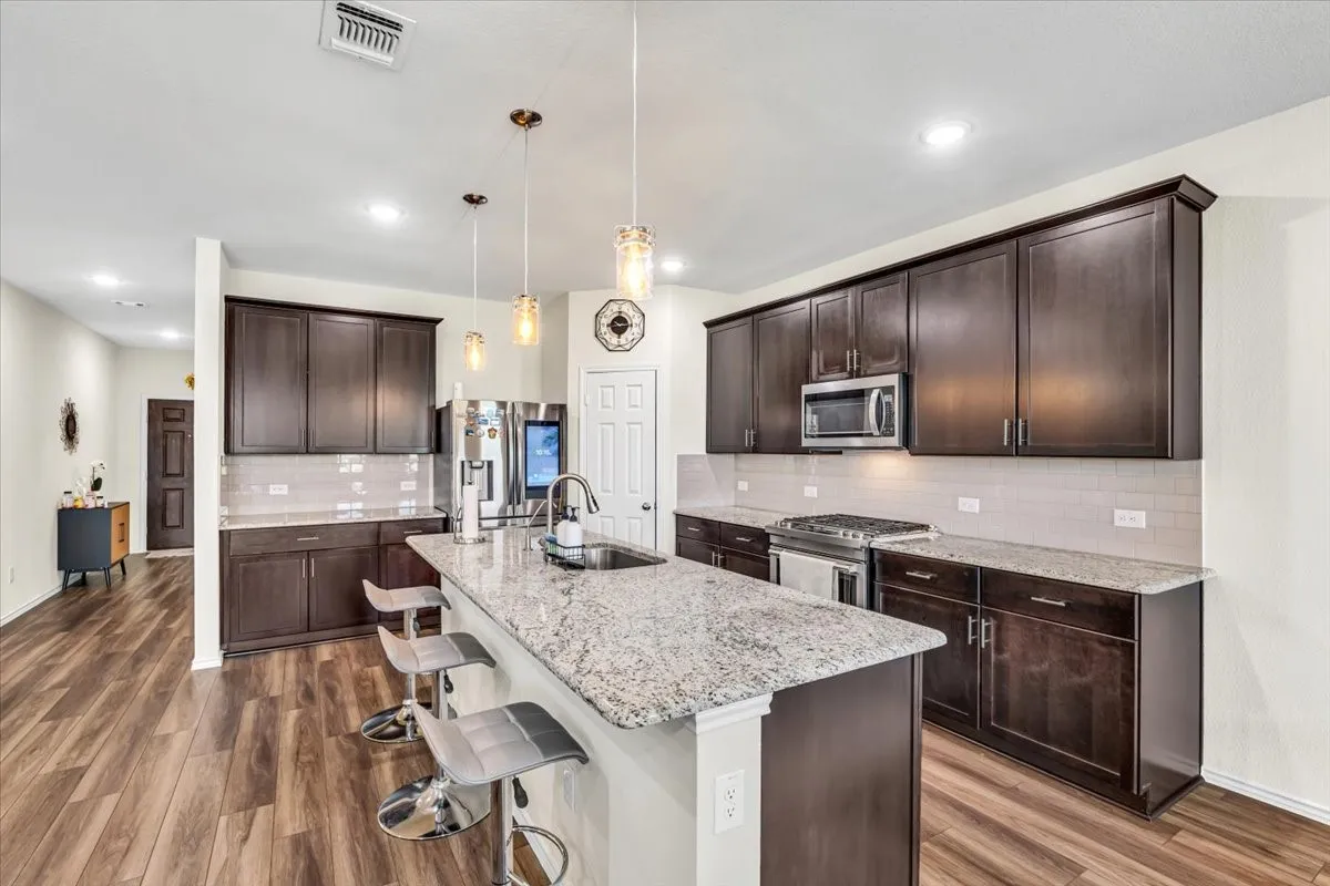 Kitchen featuring wood-finish flooring, dark wood cabinetry, granite countertops, an island with a sink, and stainless steel appliances