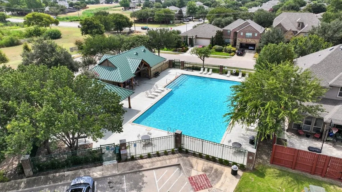 Community swimming pool with surrounding concrete decking, a covered pavilion with a green metal roof, and perimeter fencing