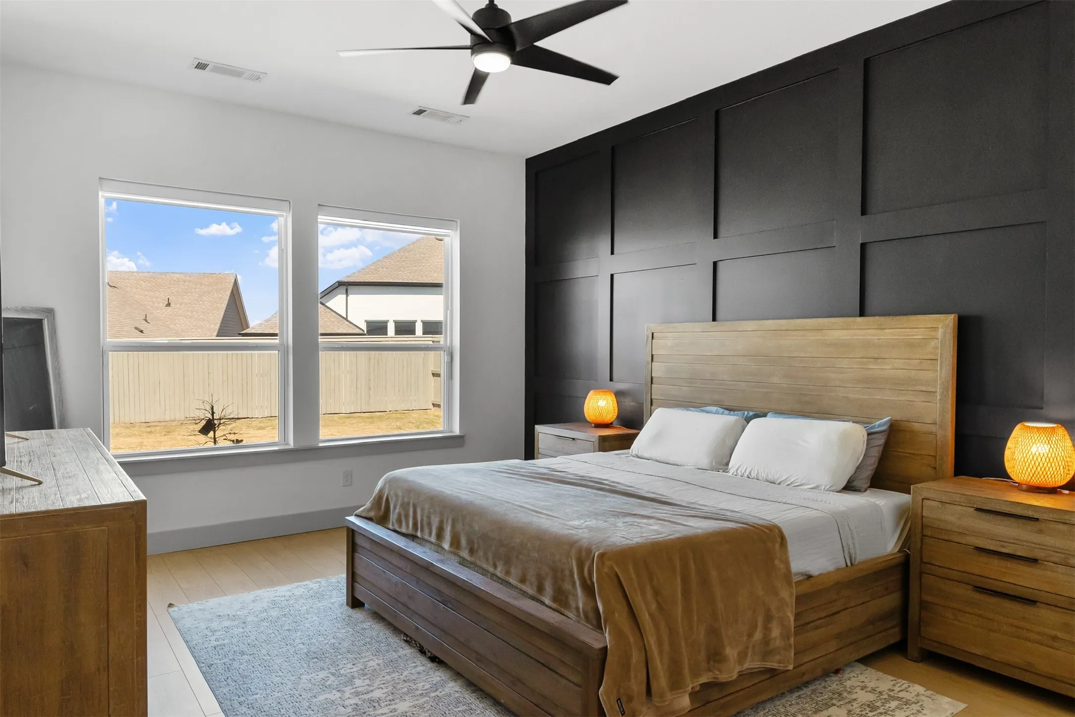 Light wood-finish flooring, accent wall with dark paneled millwork, black ceiling fan, and multiple windows providing natural illumination