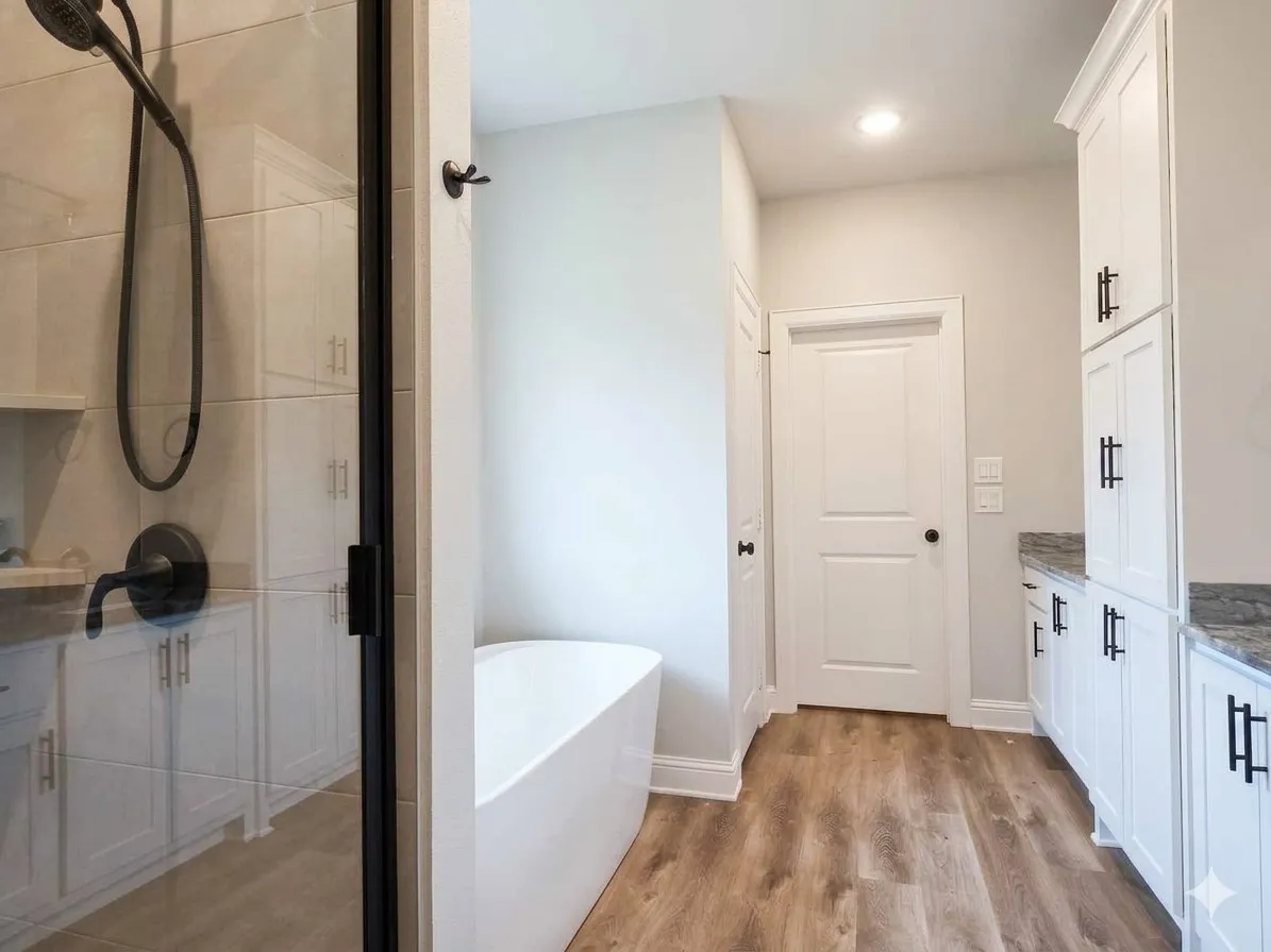 Freestanding white soaking tub, wood-finish flooring, white cabinetry with dark hardware, glass-enclosed shower with dark fixtures, and light-toned countertops