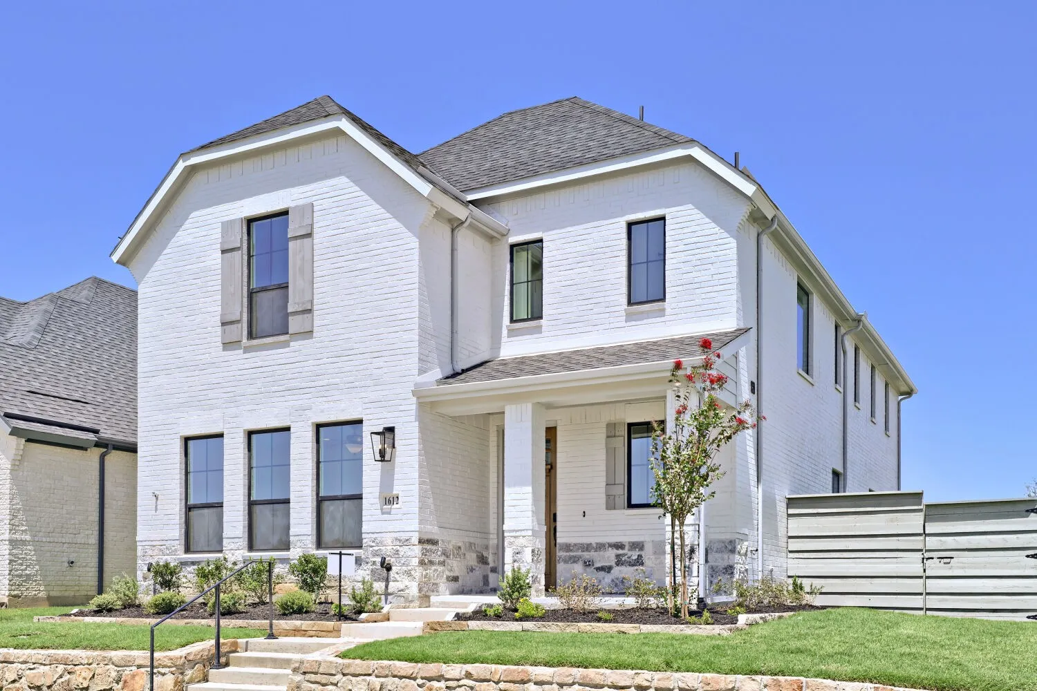 Two-story residence featuring white brick and stone veneer, dark-framed windows, a covered front porch, and mature landscaping