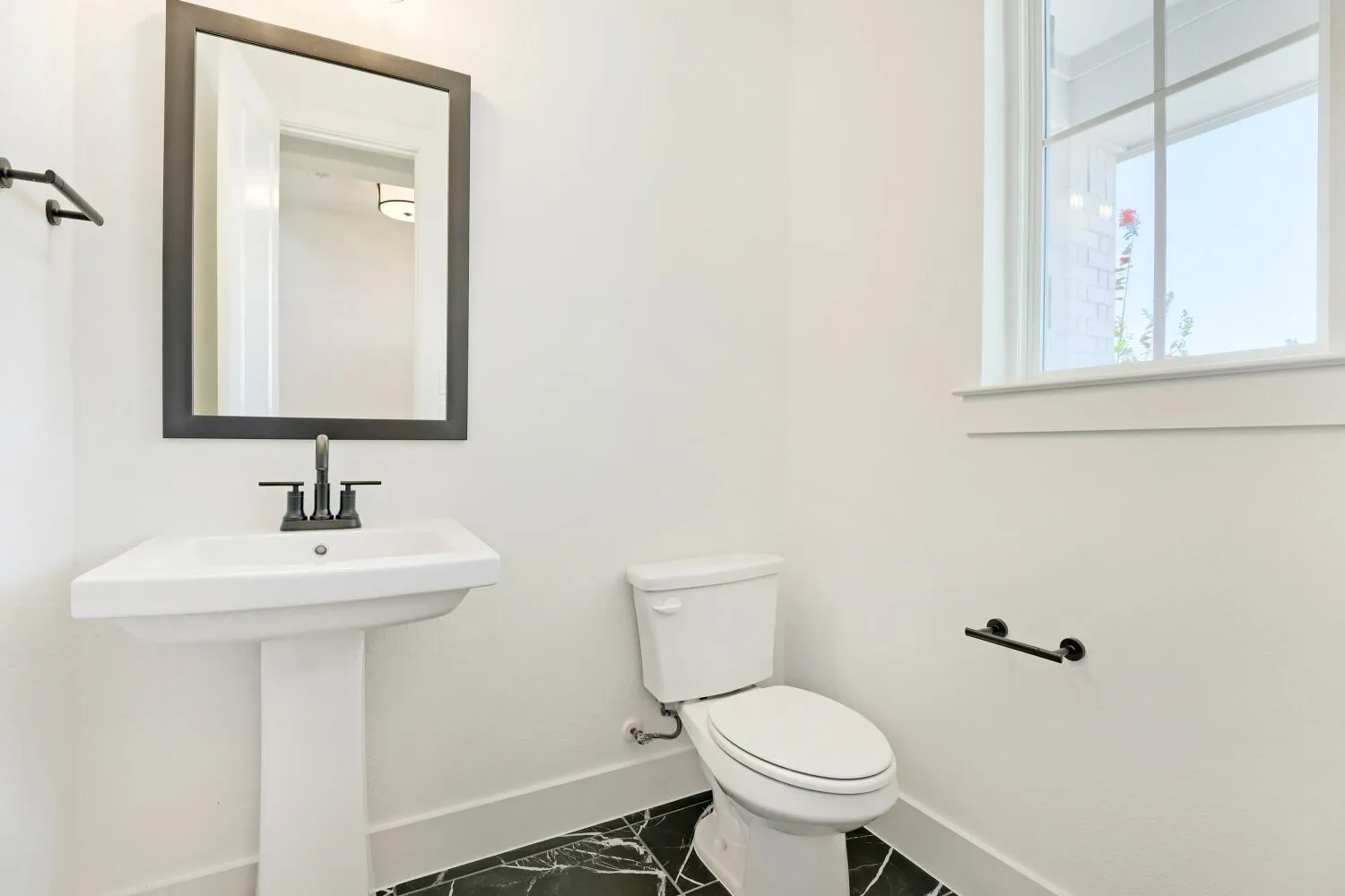 Pedestal sink with matte black faucet, framed mirror, white toilet, black and white floor tiles, and a multi-pane window