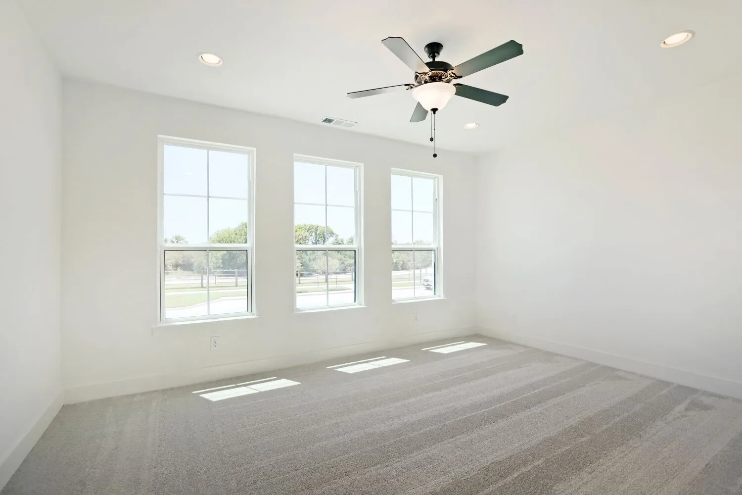 Three large windows, recessed ceiling lighting, ceiling fan with integrated light, light gray carpeting, and white walls