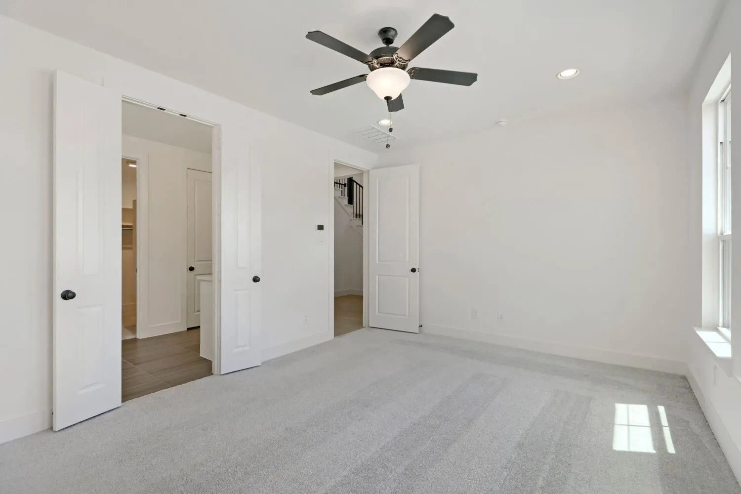Recessed lighting and a ceiling fan fixture illuminate a room with light-colored carpet flooring and multiple paneled doors