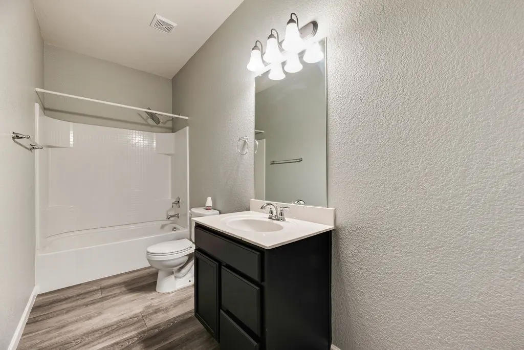 Modern bathroom featuring a black vanity with white rectangular sink and chrome fixtures, wood-finish flooring, large illuminated mirror with multi-bulb lighting, and a separate bathtub-shower combination with white subway tile surround and glass enclosure