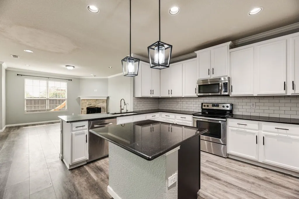 Modern kitchen featuring white cabinetry with chrome hardware, stainless steel appliances, a black quartz island countertop with waterfall edge, subway tile backsplash, recessed and pendant lighting, and wood-finish flooring throughout the open​​‌​​​​‌​​‌‌​‌‌​​​‌‌​‌​‌​‌​​​‌​​ layout