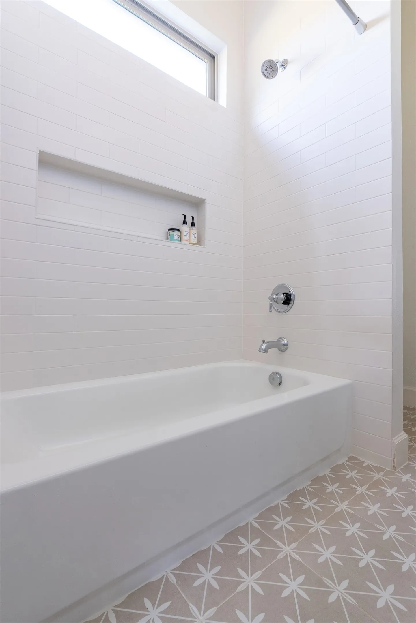 White subway tile surround, built-in shower niche, chrome plumbing fixtures, white bathtub, patterned floor tile, and transom window