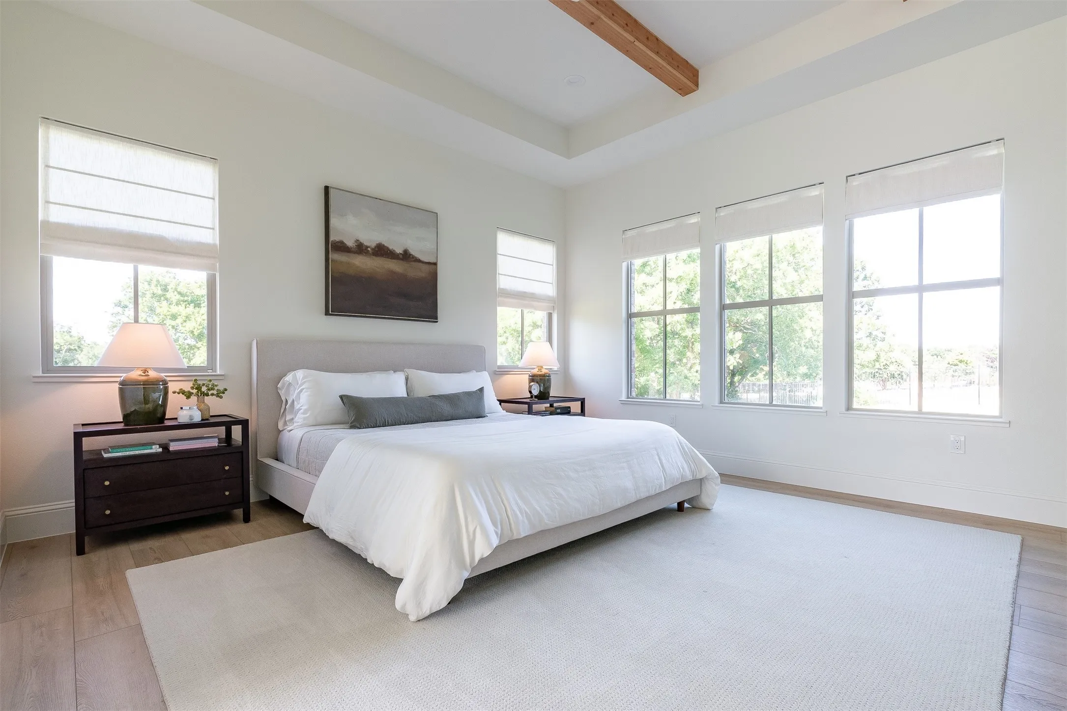 Tray ceiling with exposed wood beam, multiple large windows with Roman shades, wood-finish flooring, and white walls